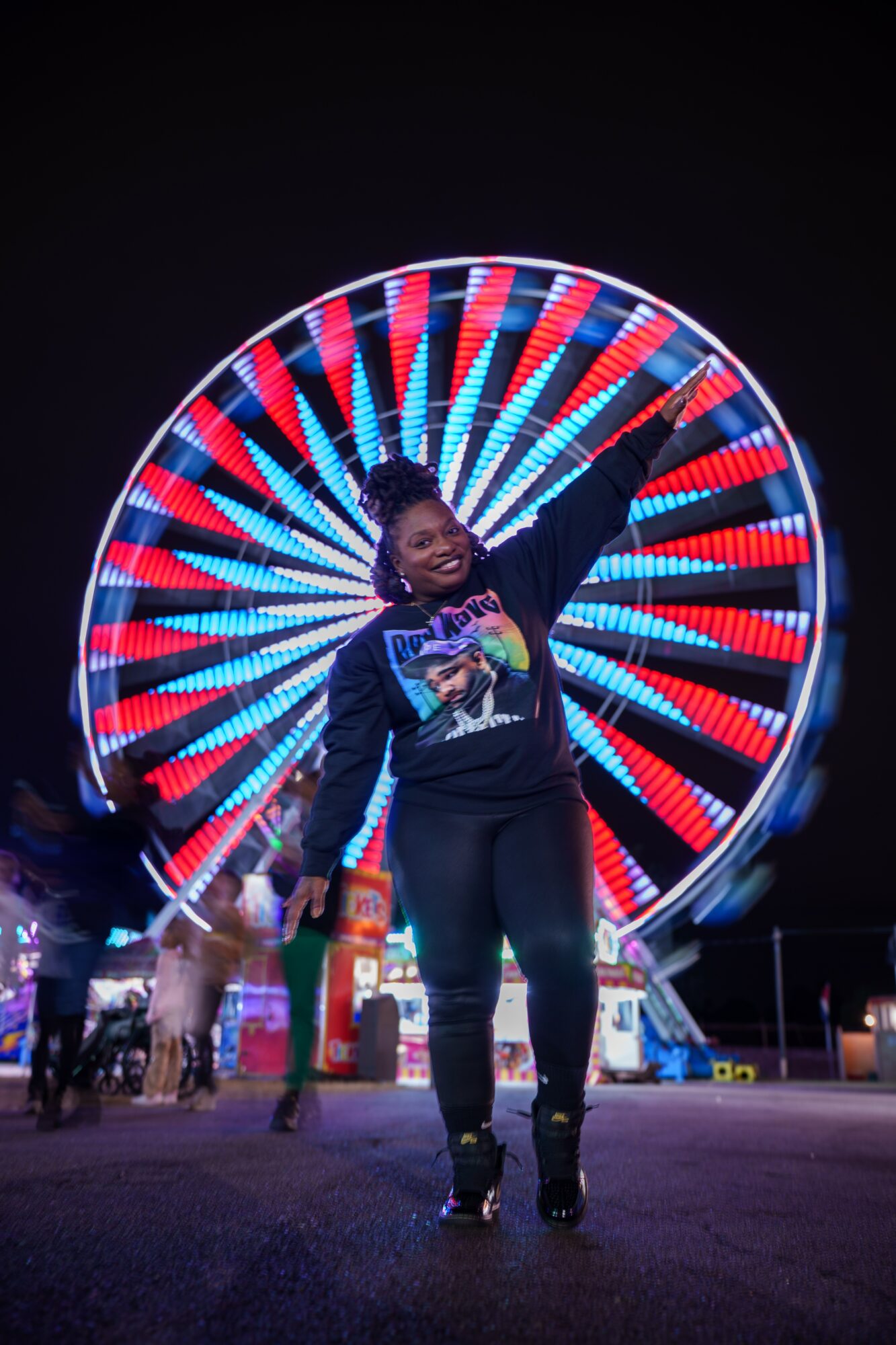 Person smiling and waving in front of a brightly lit Ferris wheel at night, with colorful lights forming a circular pattern.