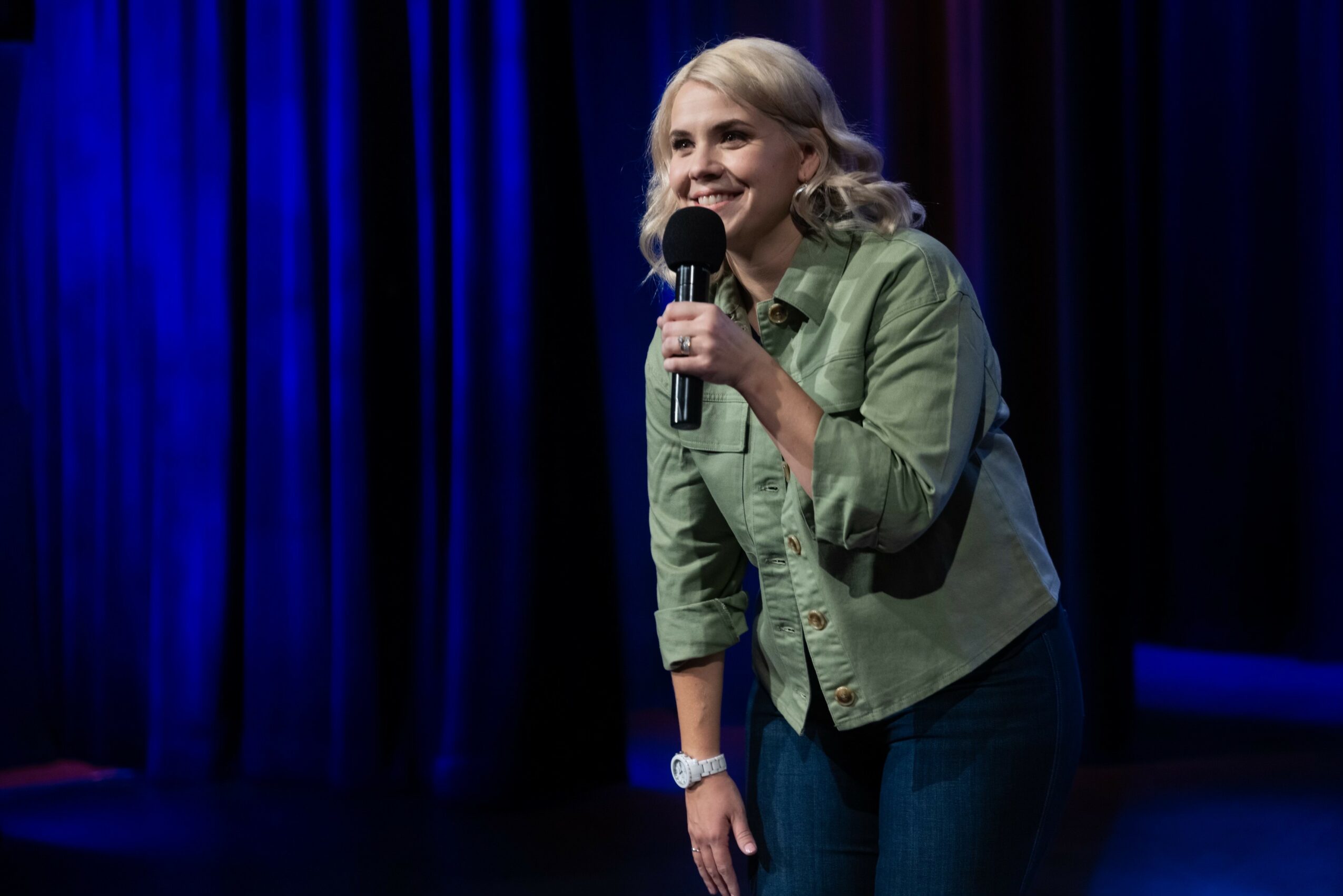 Woman with blonde hair holding a microphone on stage with blue and purple curtains behind her.
