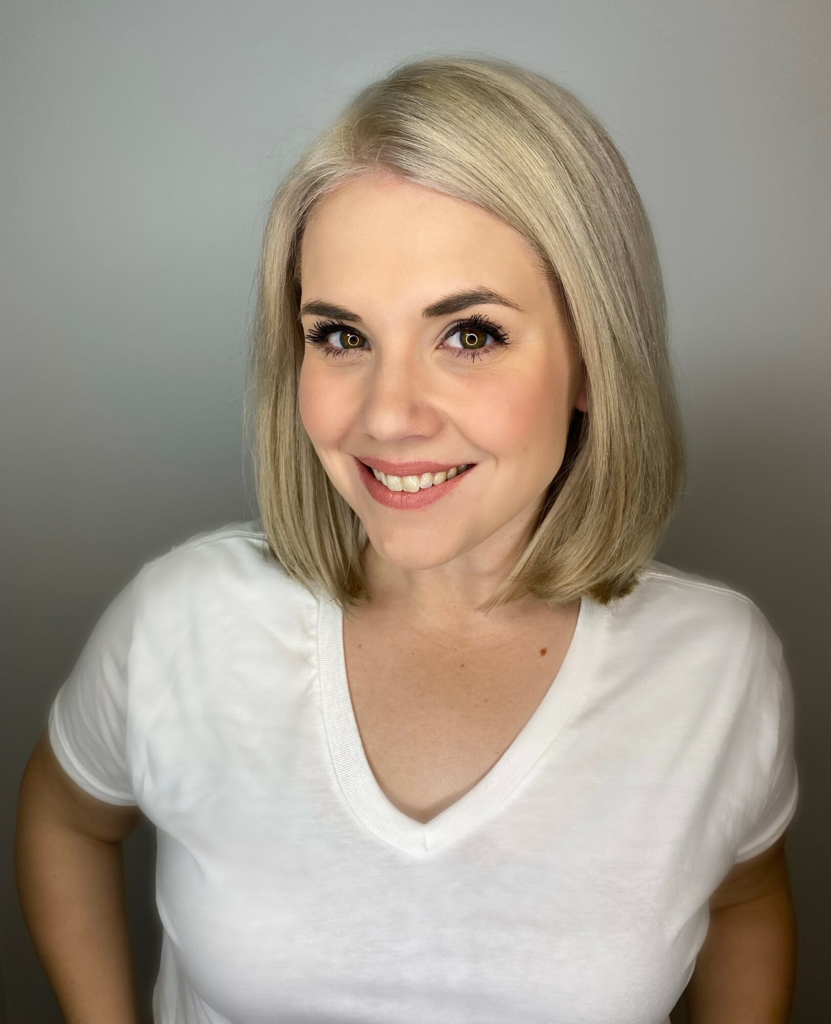 Portrait of a smiling woman with shoulder-length blonde hair, wearing a white V-neck shirt, against a neutral background.