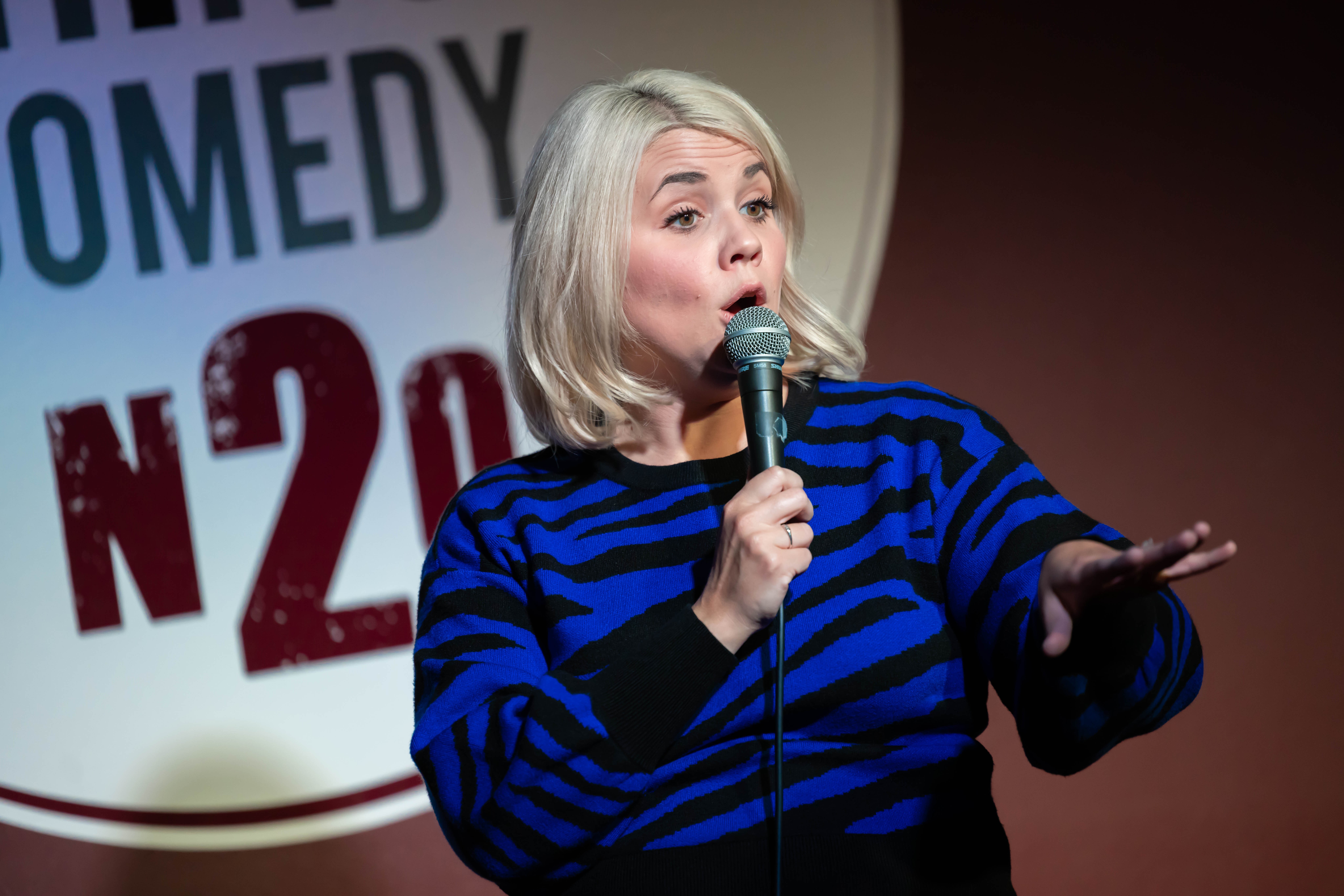 Woman with blonde hair speaking into a microphone on stage, wearing a blue and black striped top, with a comedy sign in the background.