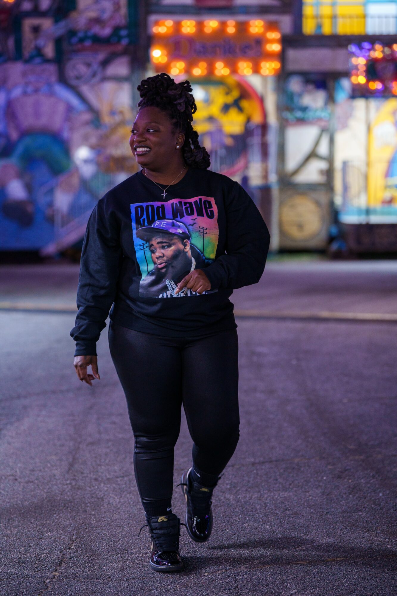 Person walking outdoors at night, wearing a black hoodie with a colorful graphic, smiling, with blurred amusement park lights in background.