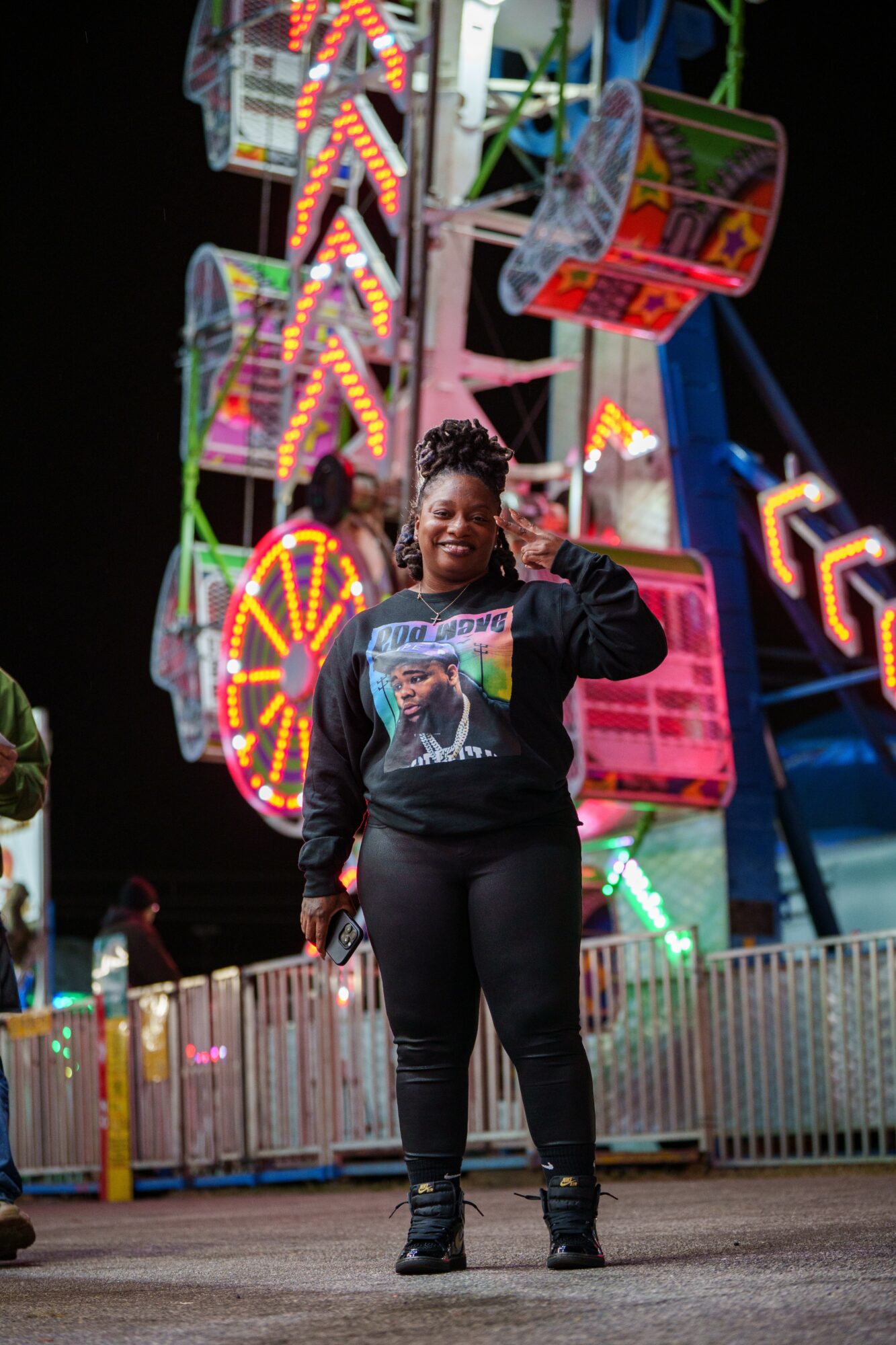 Person standing in front of a brightly lit amusement park ride at night, smiling and giving a thumbs-up gesture.