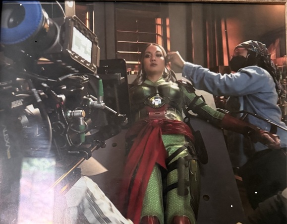 A woman in a green costume with red accents is being prepared for filming, with a crew member adjusting her hair.
