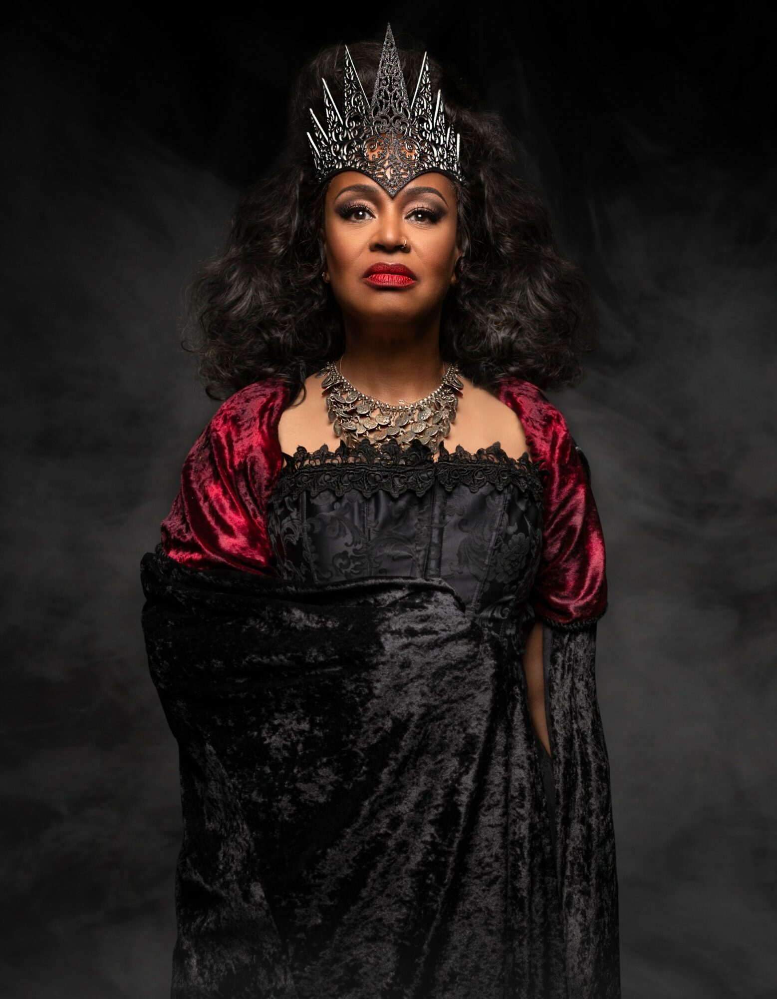 Woman wearing a crown, red and black dress, and jewelry, standing against a dark background.