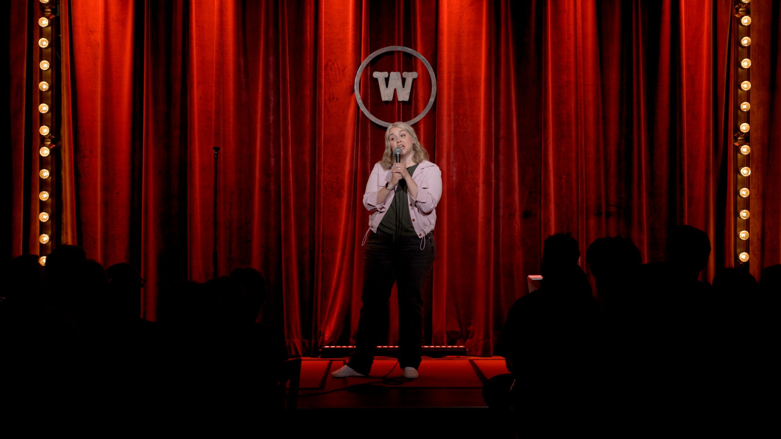 Person standing on stage with red curtains, holding a microphone, performing in front of an audience.