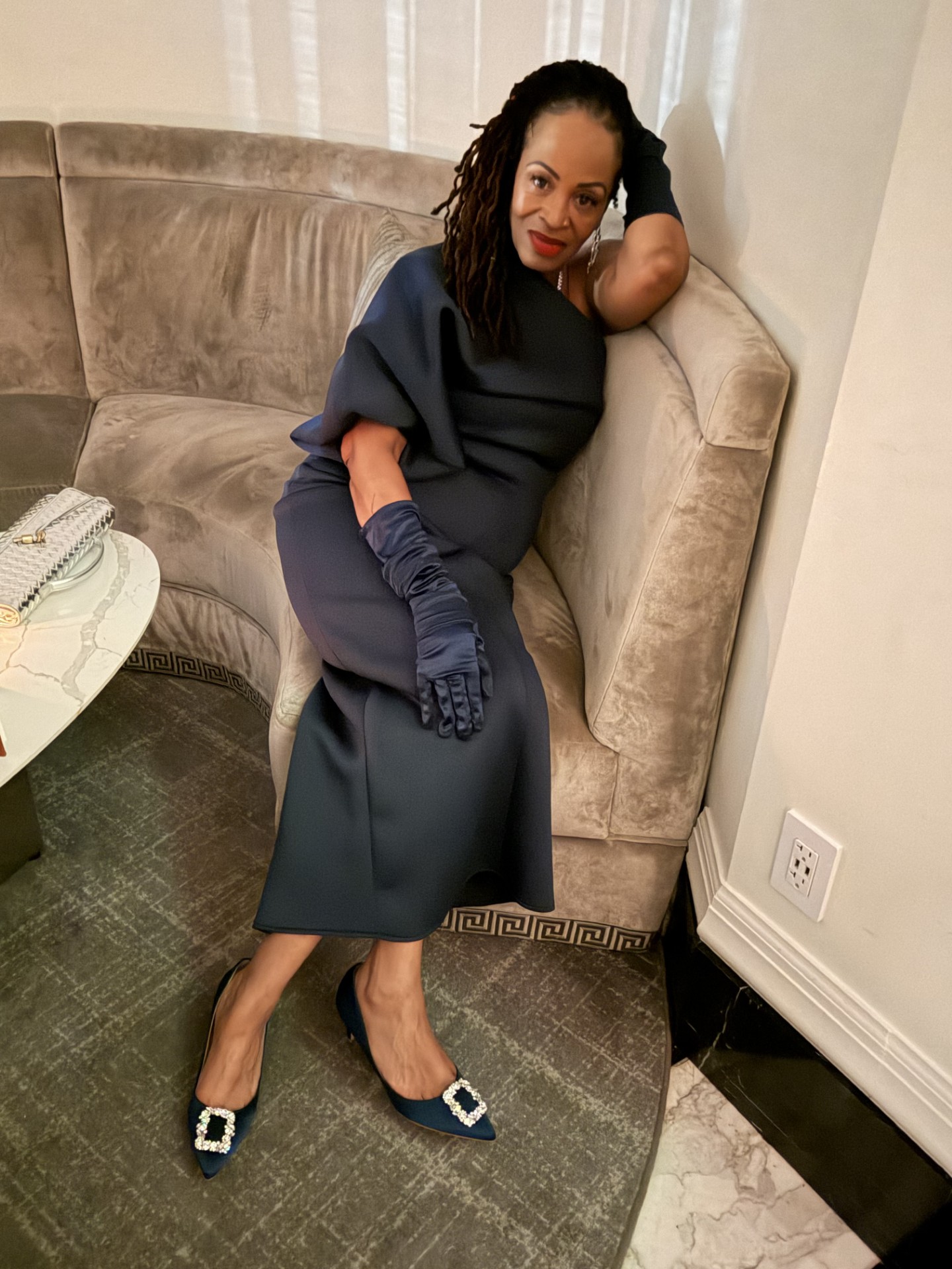 Woman sitting on a beige sofa, wearing a dark dress and gloves, with one arm resting on the sofa's back.