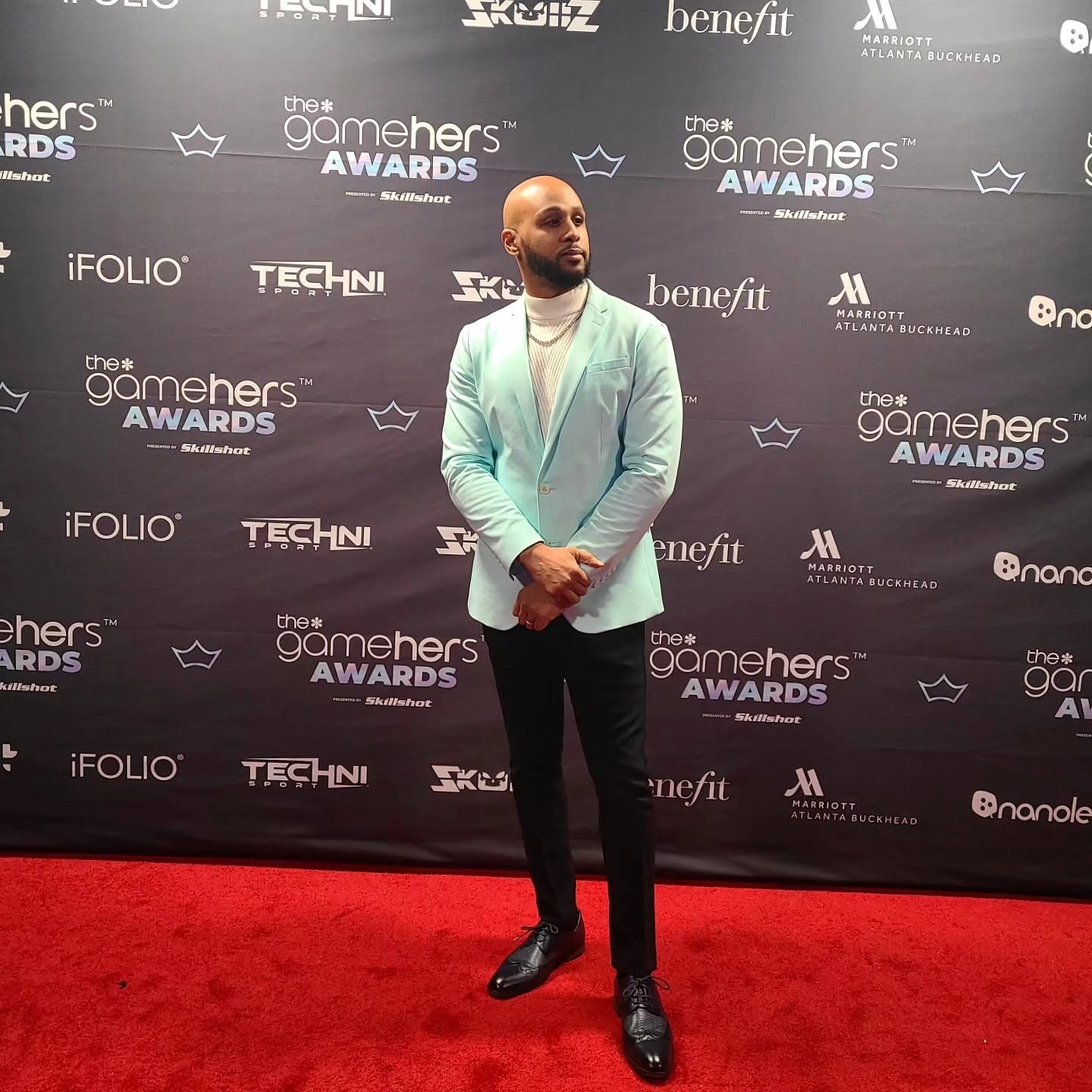 Man in light green blazer and black pants standing on red carpet at awards event, backdrop with logos behind him.