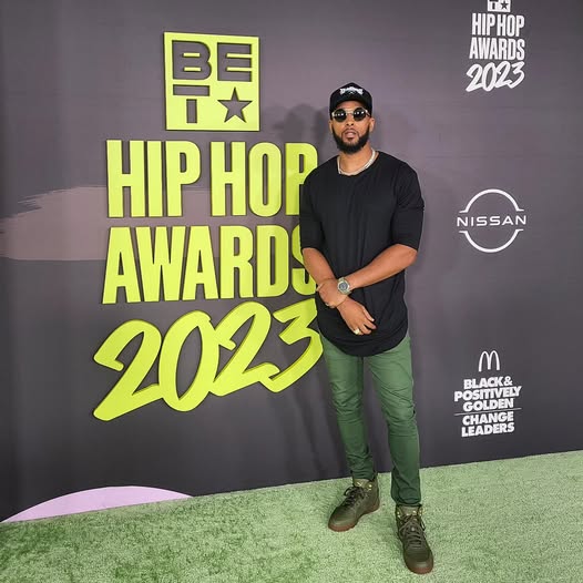 Man in black shirt and green pants standing on green carpet at awards event, backdrop with text and logos.