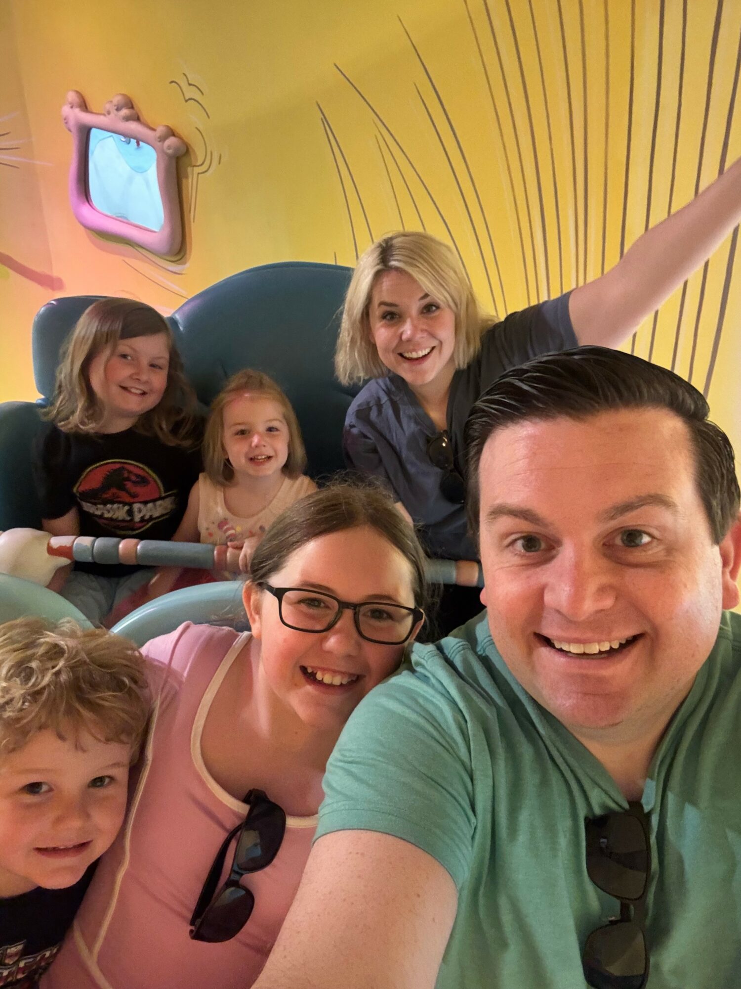 Group of five smiling people, including children and adults, taking a selfie indoors with a yellow background and a TV screen.