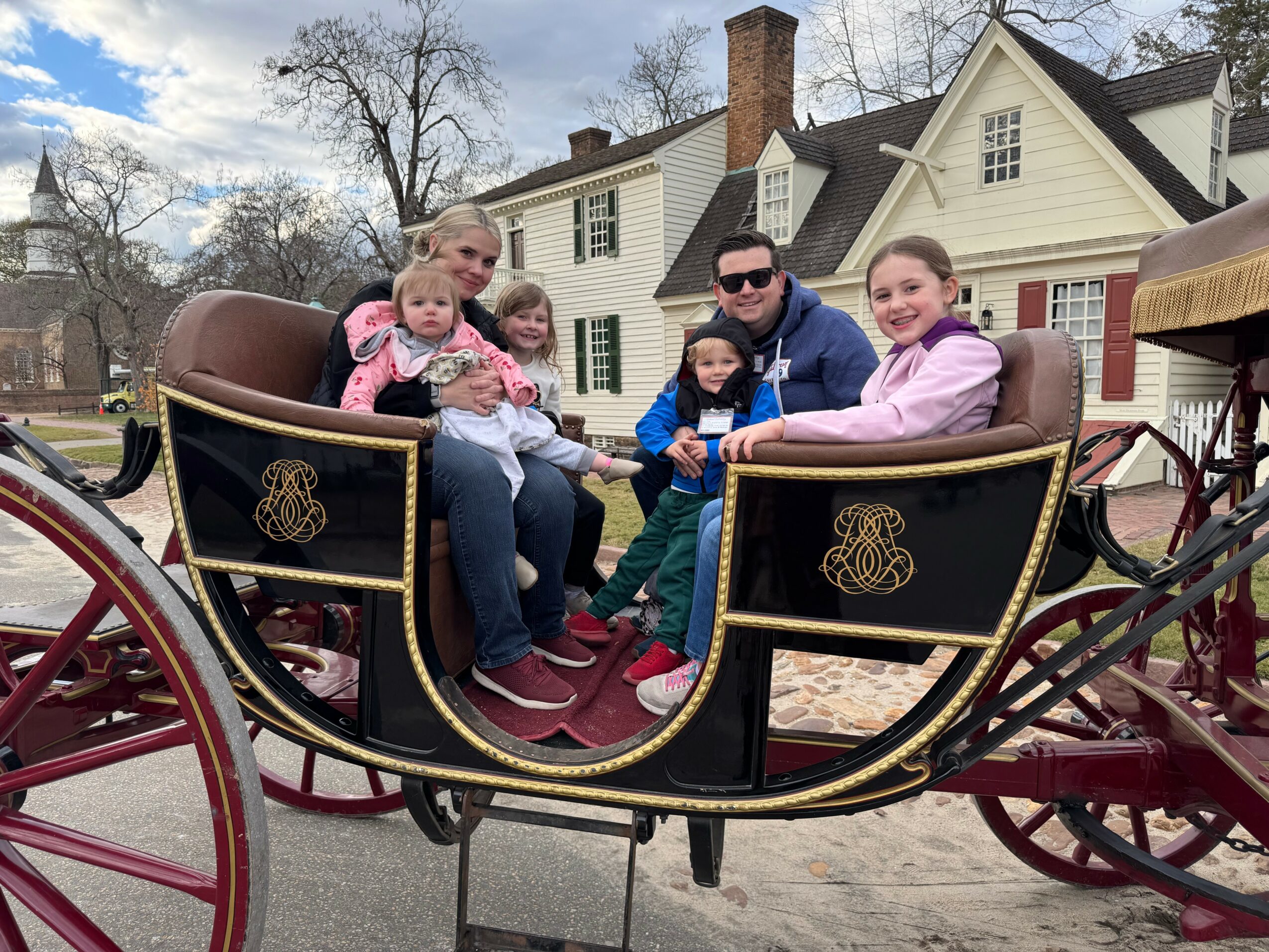 Group of six people sitting in a horse-drawn carriage outdoors, with houses and trees in the background.