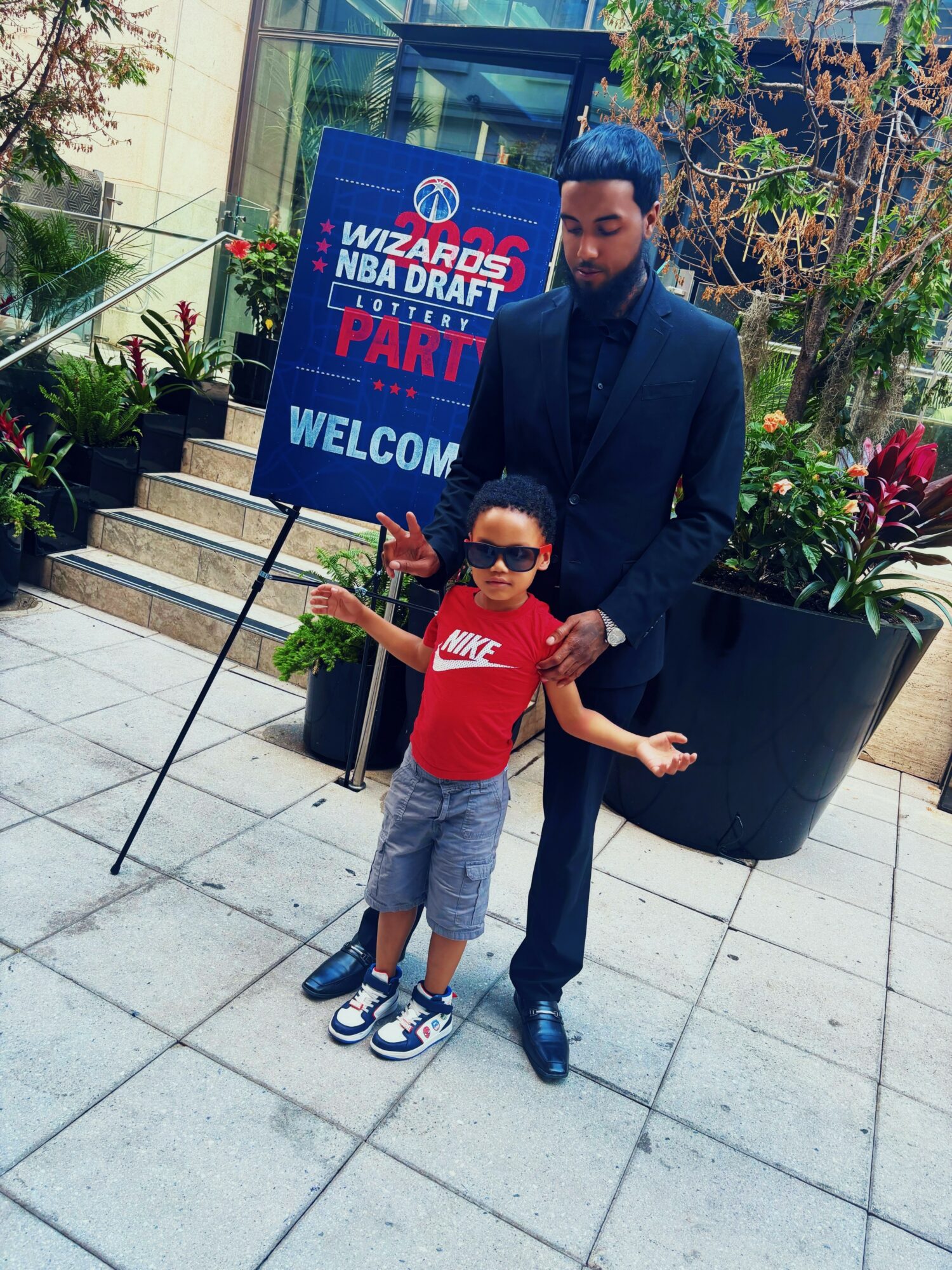 Man and child standing outside near plants, with a sign for NBA draft party in the background.