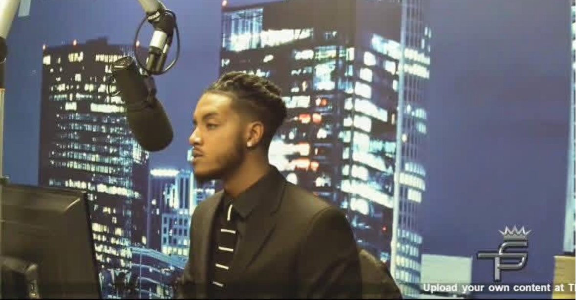 Man in suit speaking into a microphone in a recording studio with cityscape background at night.