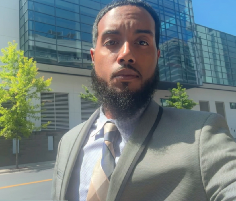 Man in suit and tie standing outdoors with modern glass building and trees in background.