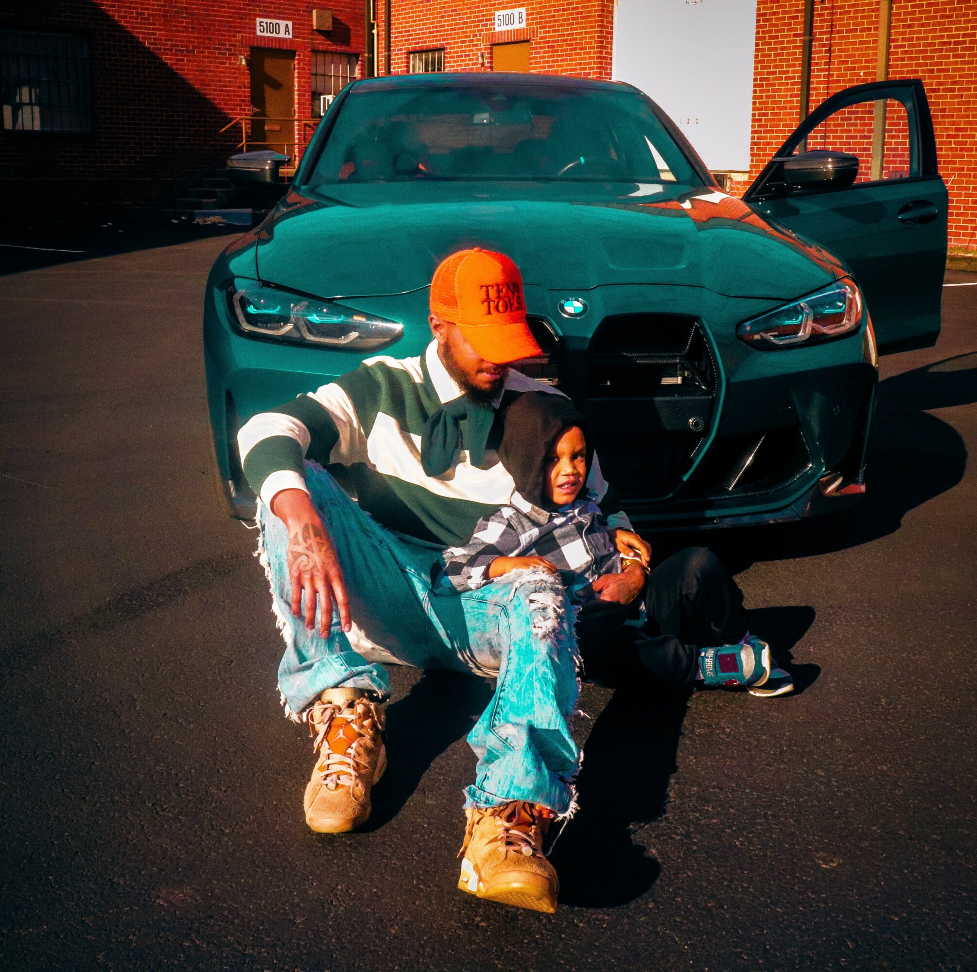 Two people sitting on the ground in front of a green car, one wearing an orange cap and the other in a striped shirt.