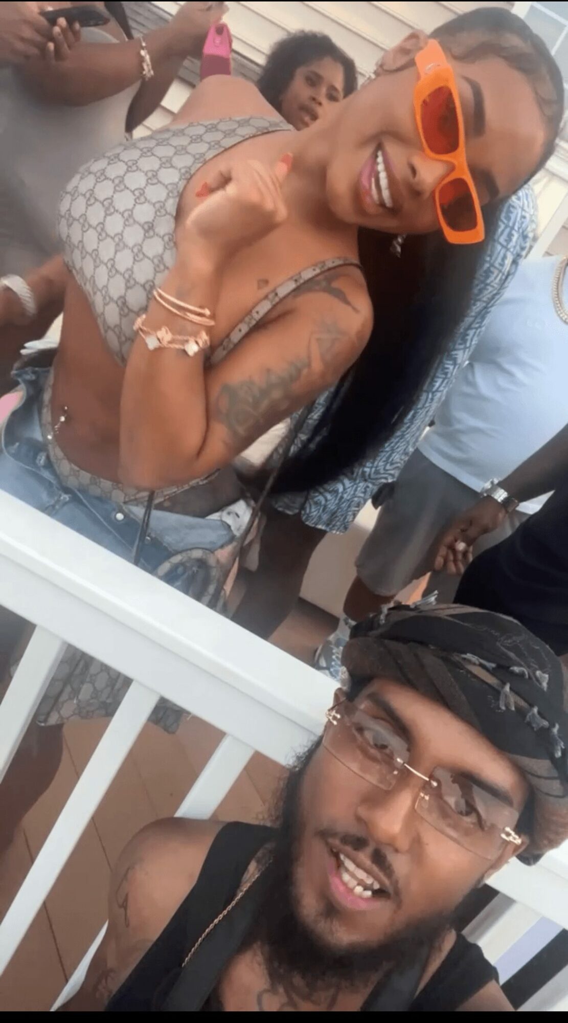 Two people smiling, one wearing orange sunglasses and a patterned top, the other with glasses and a headscarf, in a social setting.