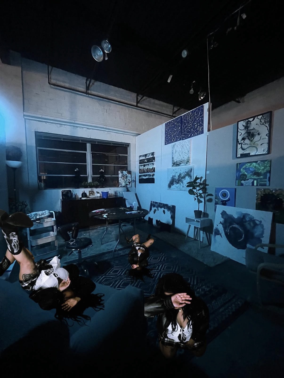 Dark room with people lying on sofas, artwork on walls, and a window with blinds, illuminated by blue lighting.