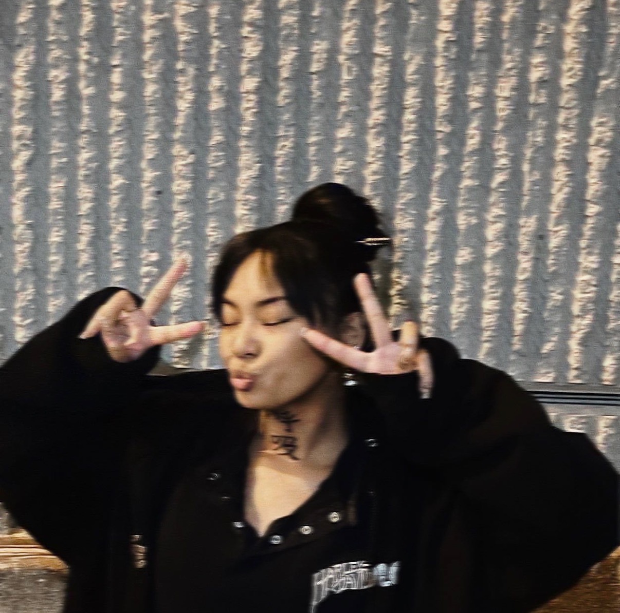 Young woman with dark hair making peace signs near her face, eyes closed, in front of textured curtain background.