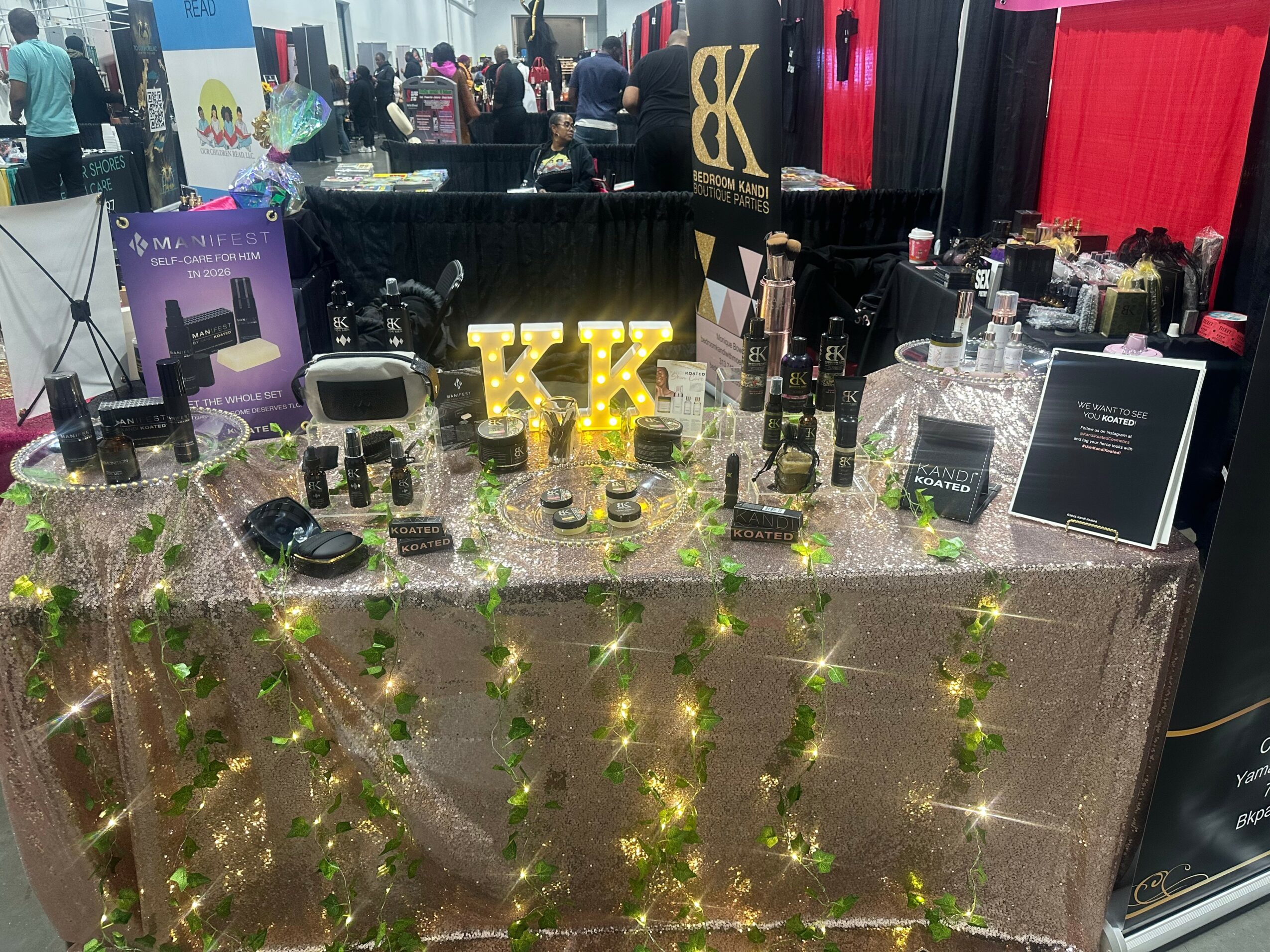 Display table with makeup products, illuminated 'KK' sign, and decorative lights, at an indoor event or market.