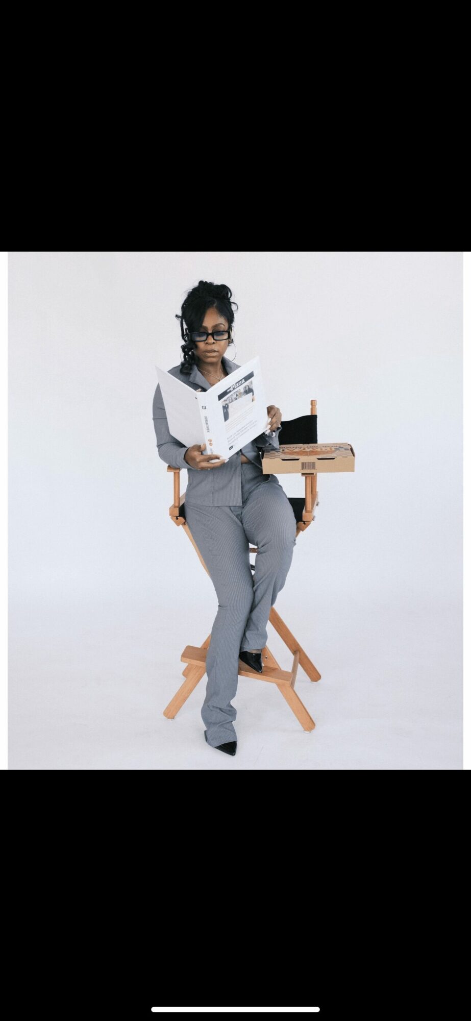 Person standing with a book, wearing glasses, gray pants, and a black top, with a wooden desk behind them.