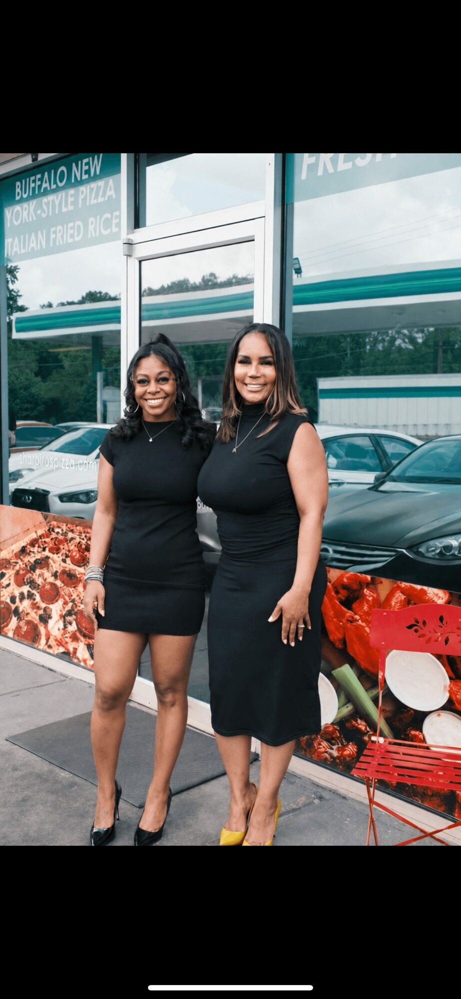 Two women stand outside a building with large glass windows, smiling, with cars parked behind them, and a red bench nearby.