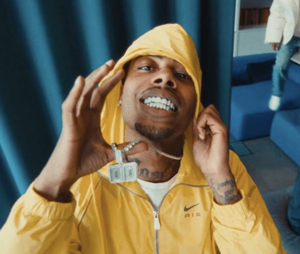Man wearing yellow hoodie and cap, smiling and gesturing with hand, indoors with blue curtains and furniture