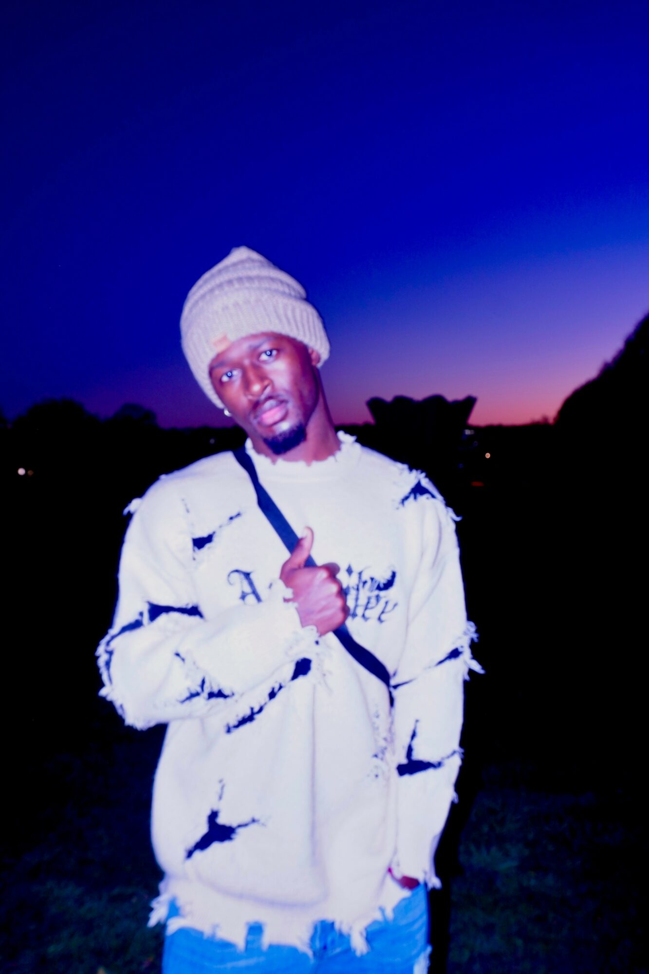 Young man wearing a white beanie and distressed jacket, standing outdoors during twilight, holding a phone, with a dark landscape background.