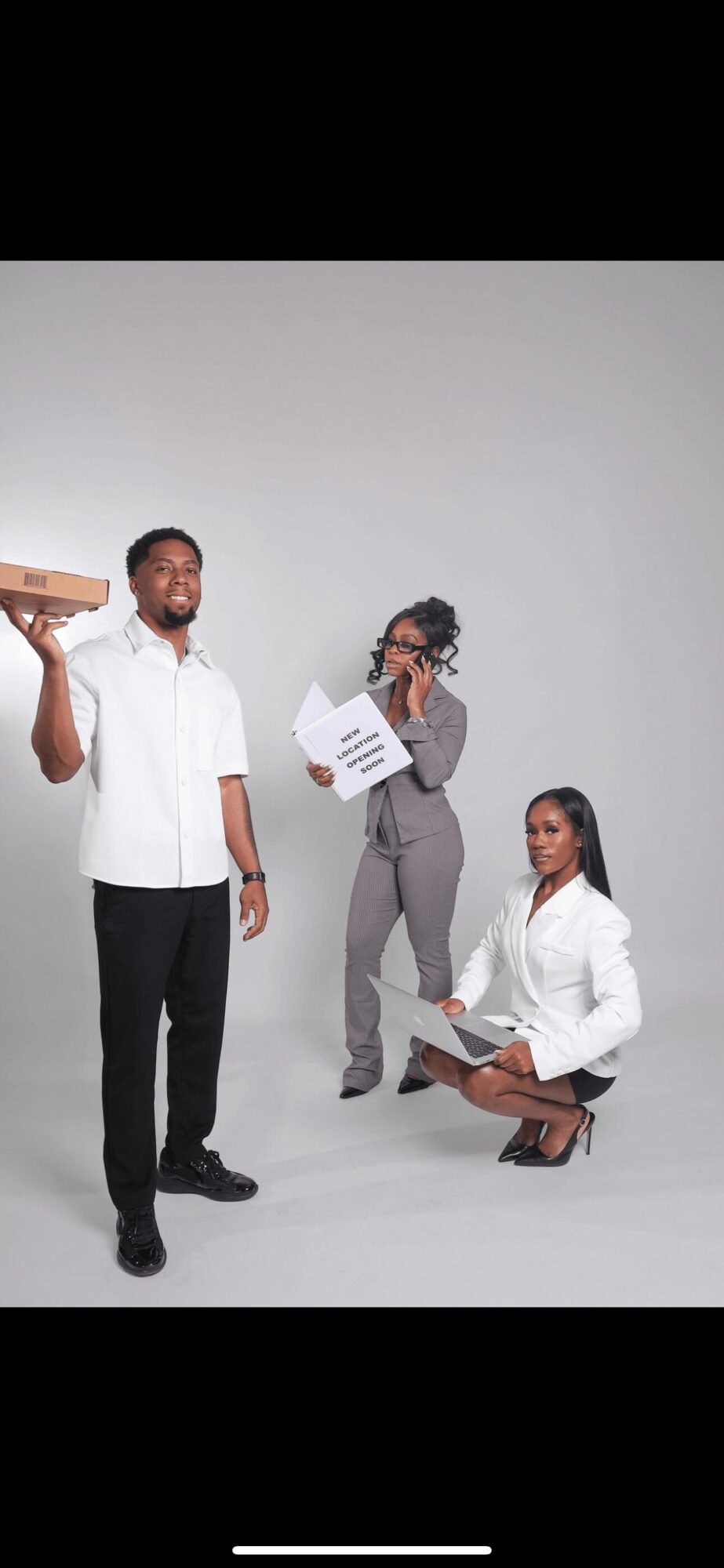 Three people in professional attire with documents and a tray, standing and sitting against a plain background.