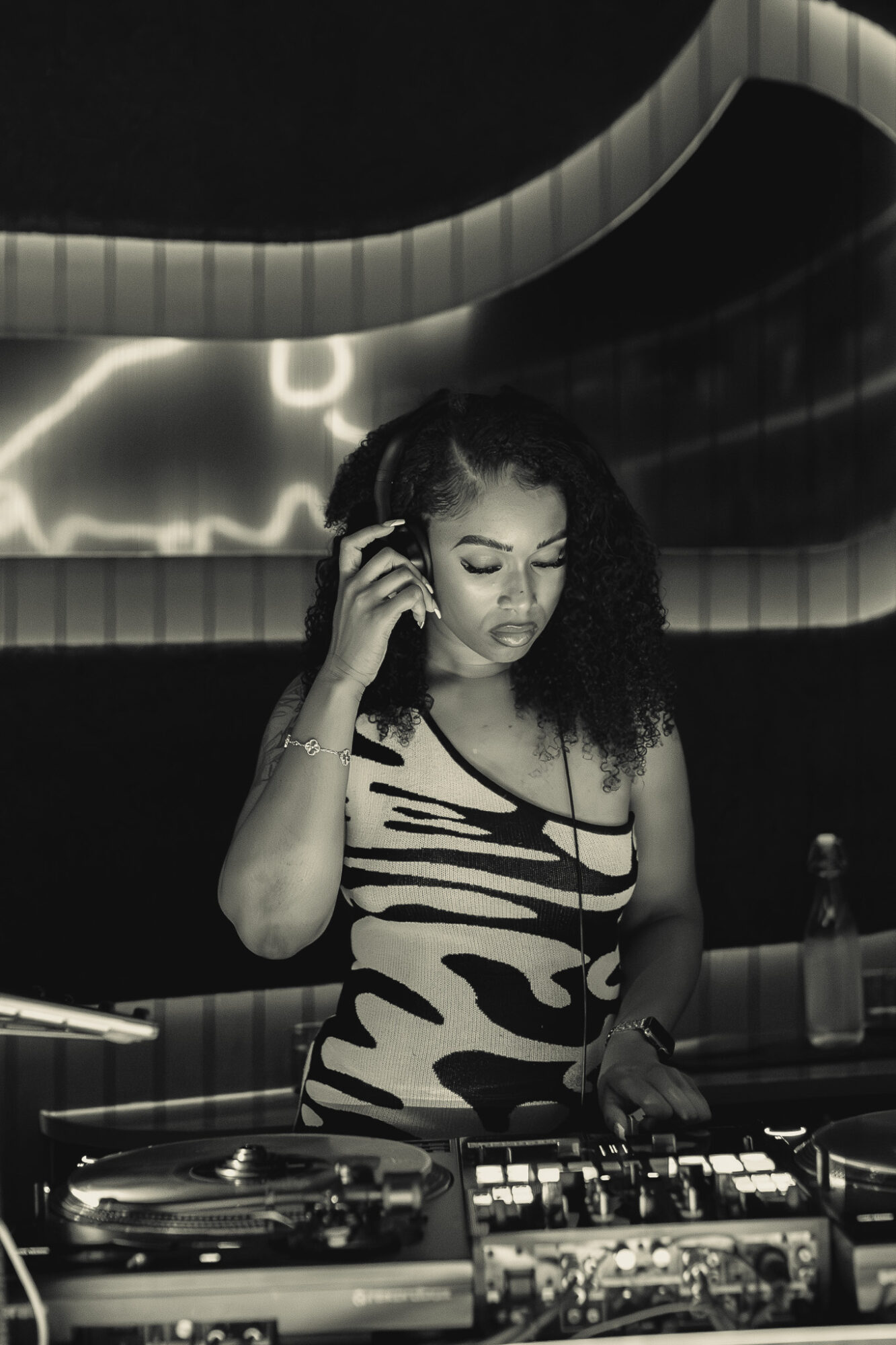 Woman with curly hair wearing a patterned dress, standing behind DJ equipment, in a dark setting with neon lights