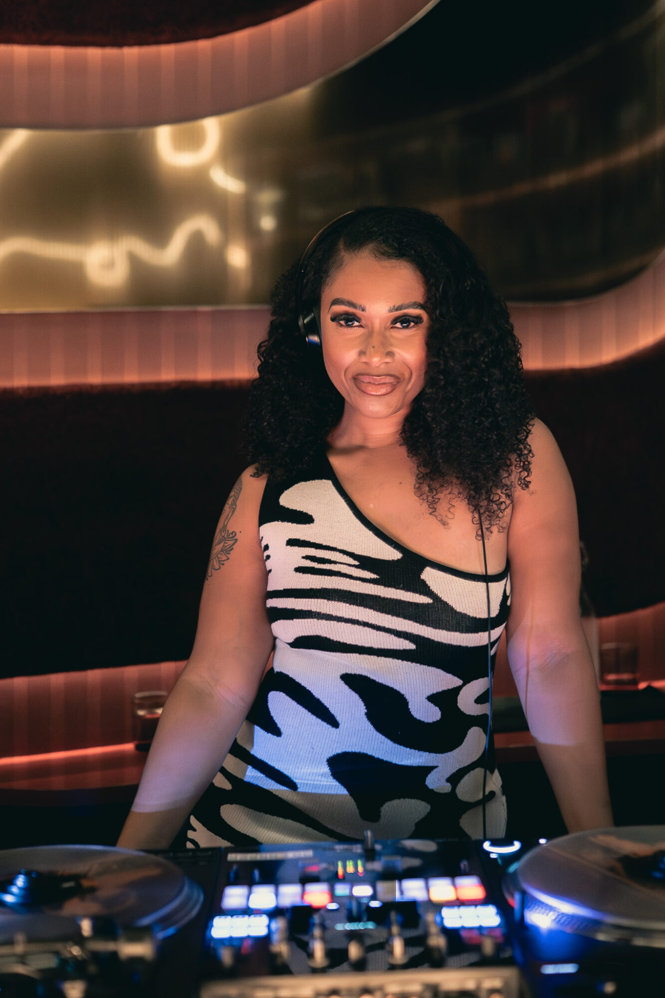 Woman with curly hair smiling, wearing a black and white patterned dress, standing in a dimly lit environment.