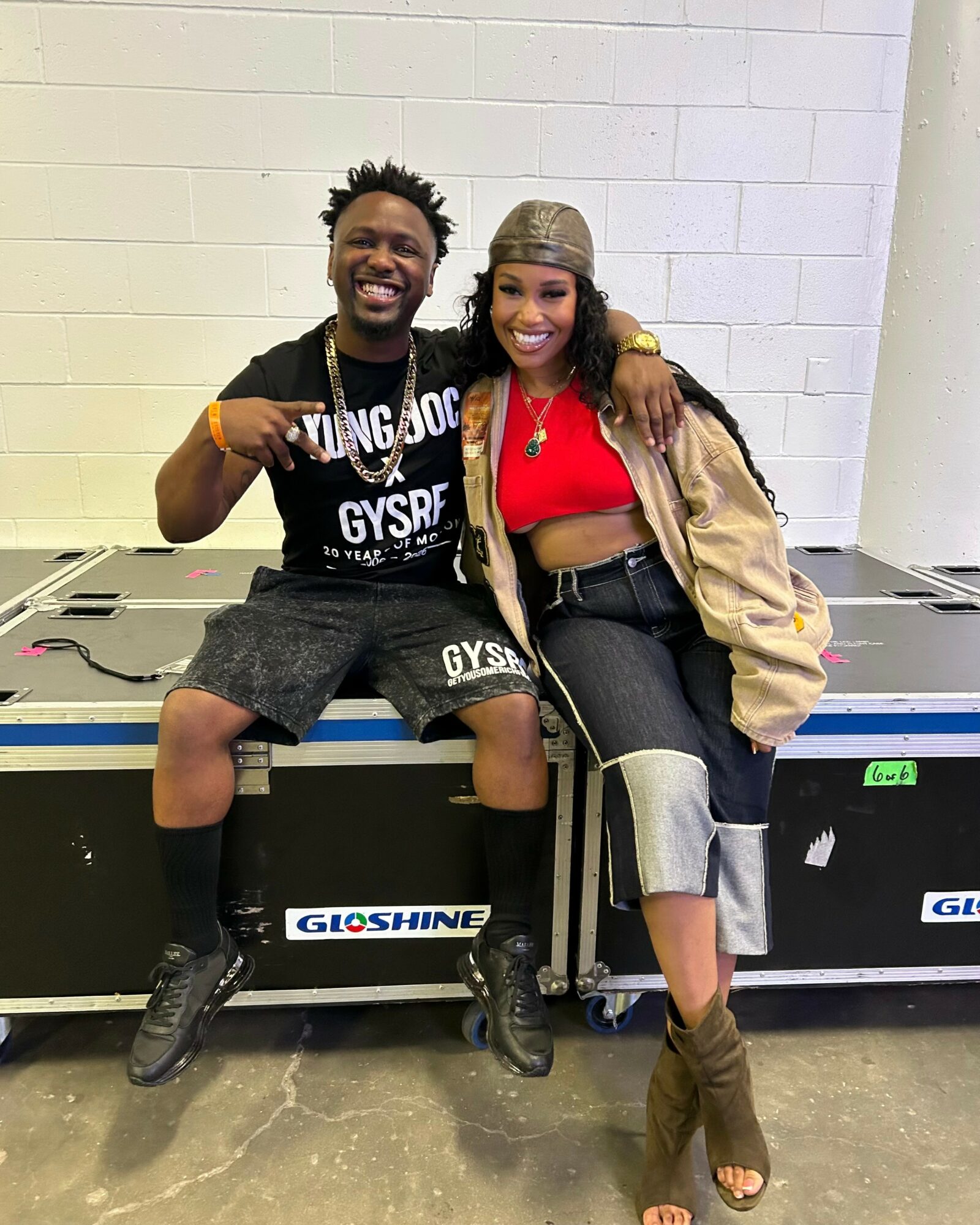 Two smiling people sit on a large black equipment case, with one person giving a thumbs-up and the other with arm around.