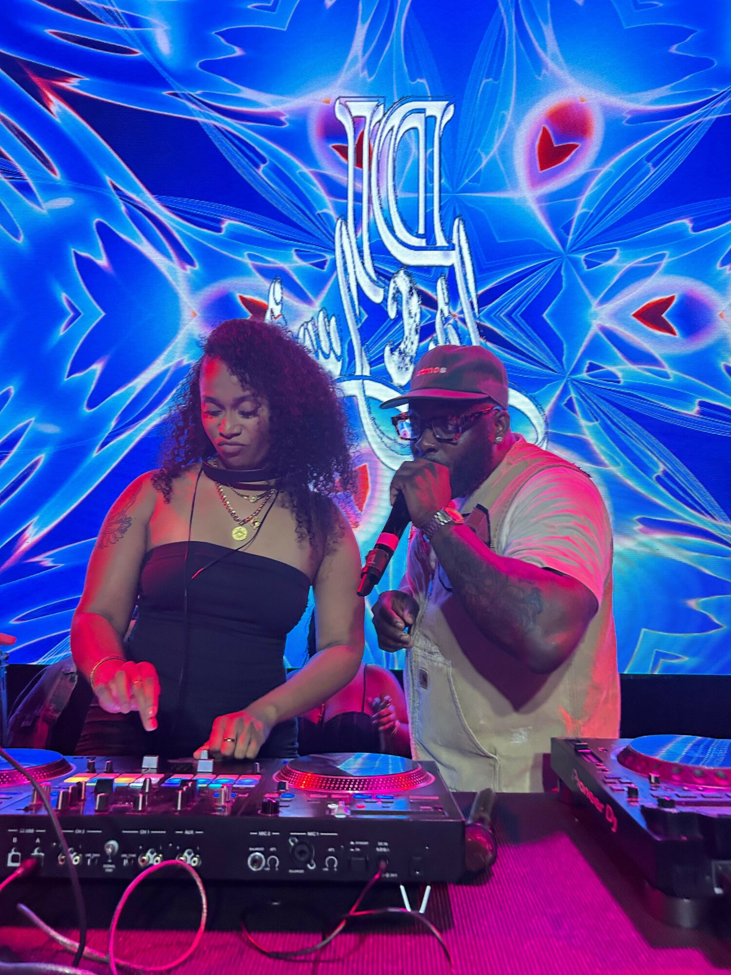 Two women DJing at a club with a vibrant blue and pink neon background, one wearing a white top and the other a black top.