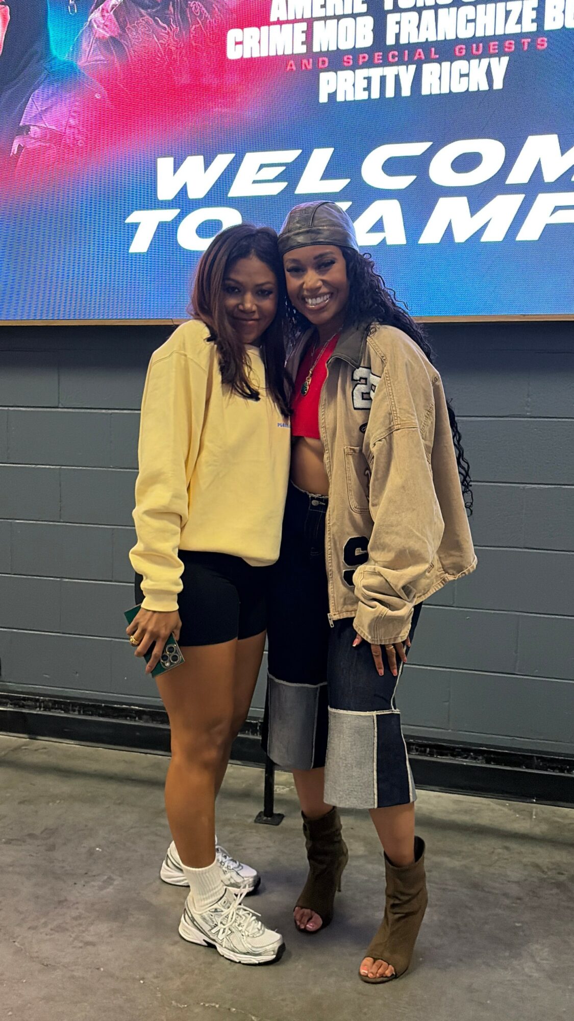 Two women standing together in front of a large screen, smiling, one in a yellow jacket and shorts, the other in a beige jacket and jeans.