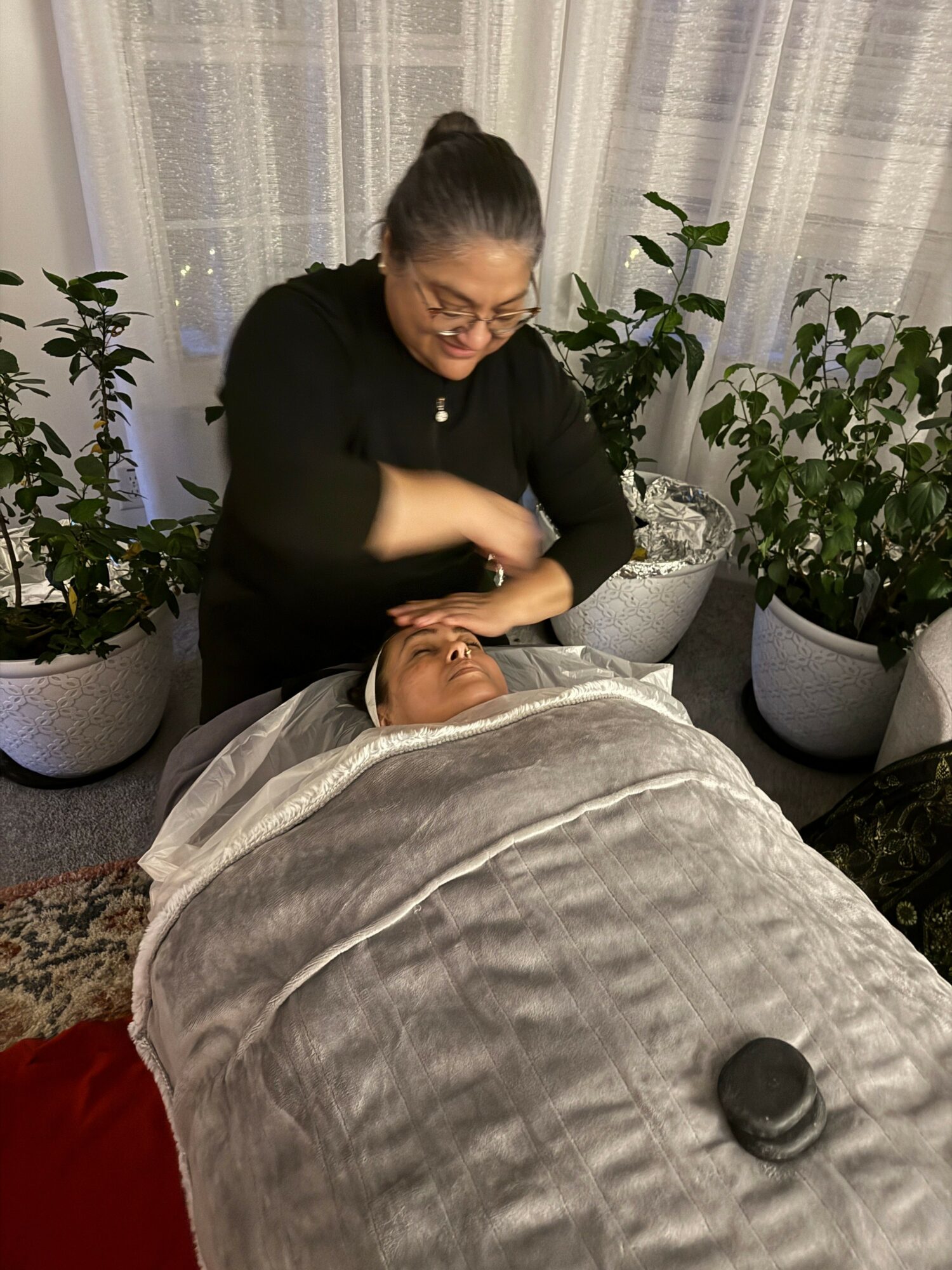 Person receiving a massage while lying on a massage table, with a therapist attending to their face, surrounded by plants.