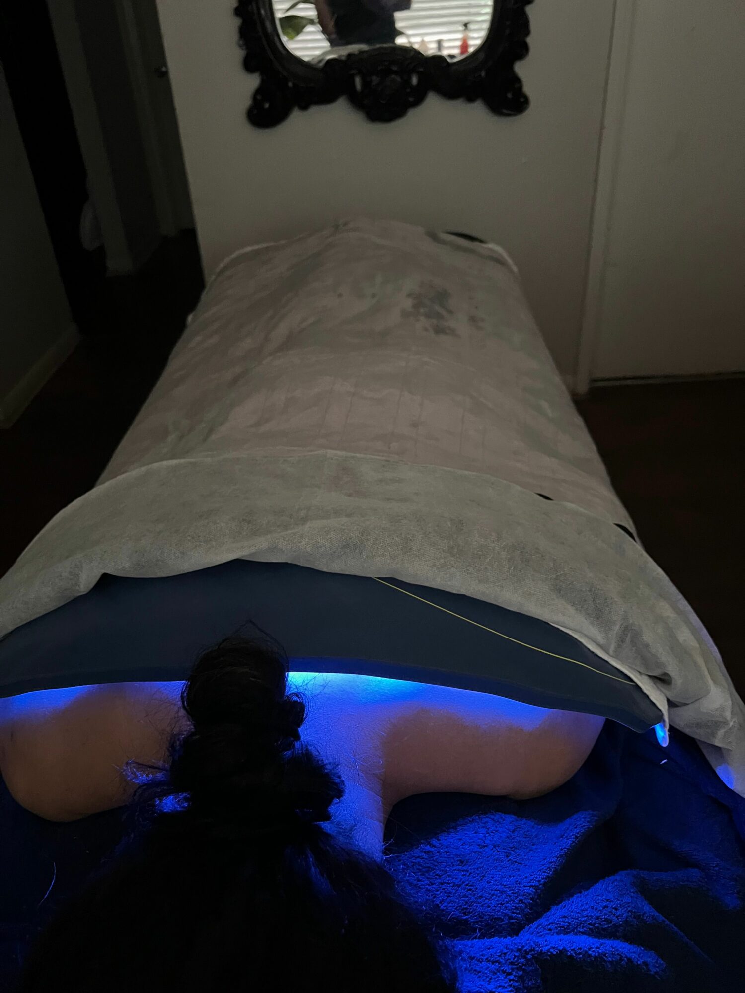 Person lying in bed with a blue light, head turned away, dark hair, and a mirror on the wall nearby.