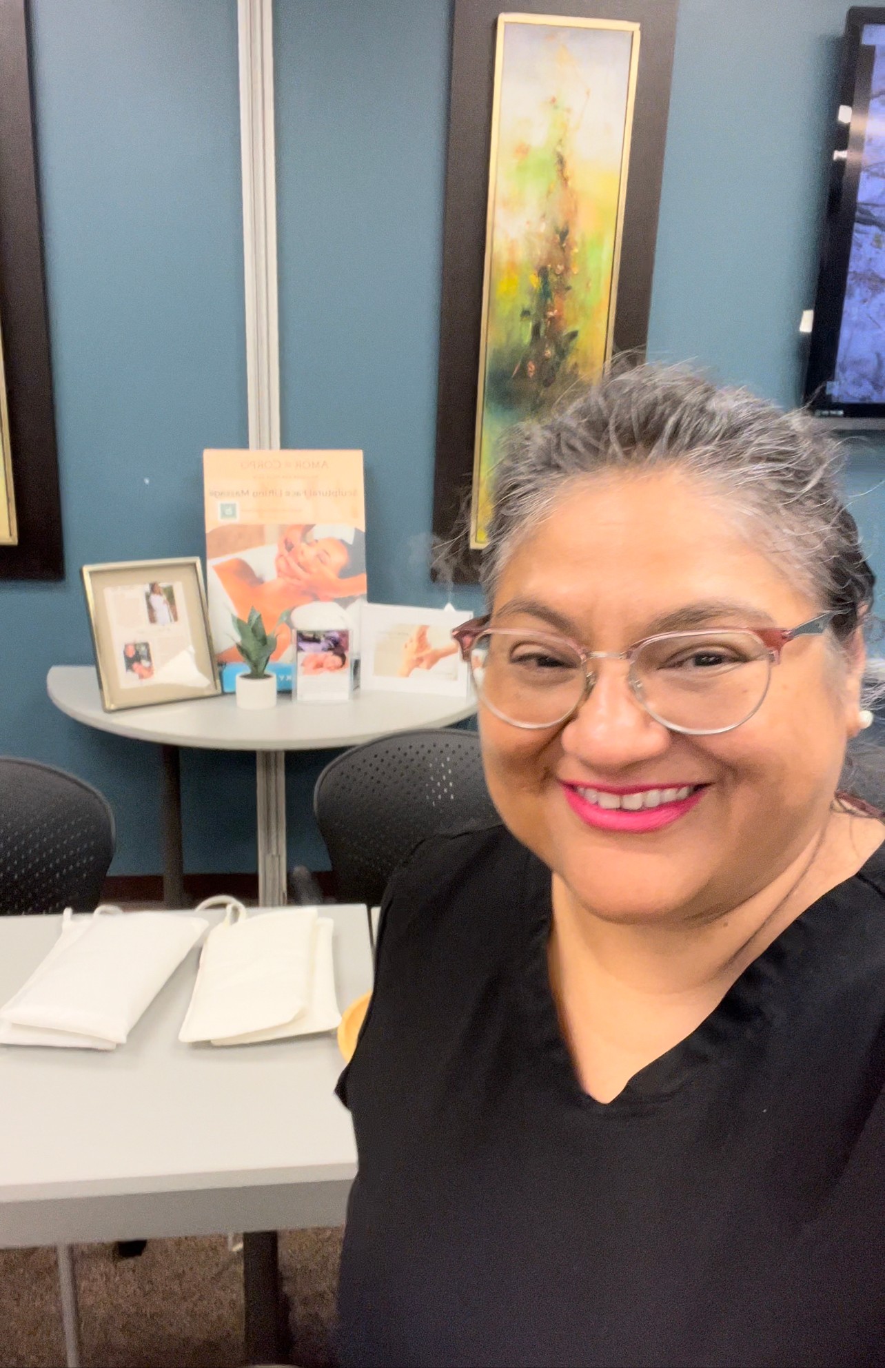 Smiling woman with glasses and gray hair in a black top, in an office or classroom setting with artwork and supplies behind her.
