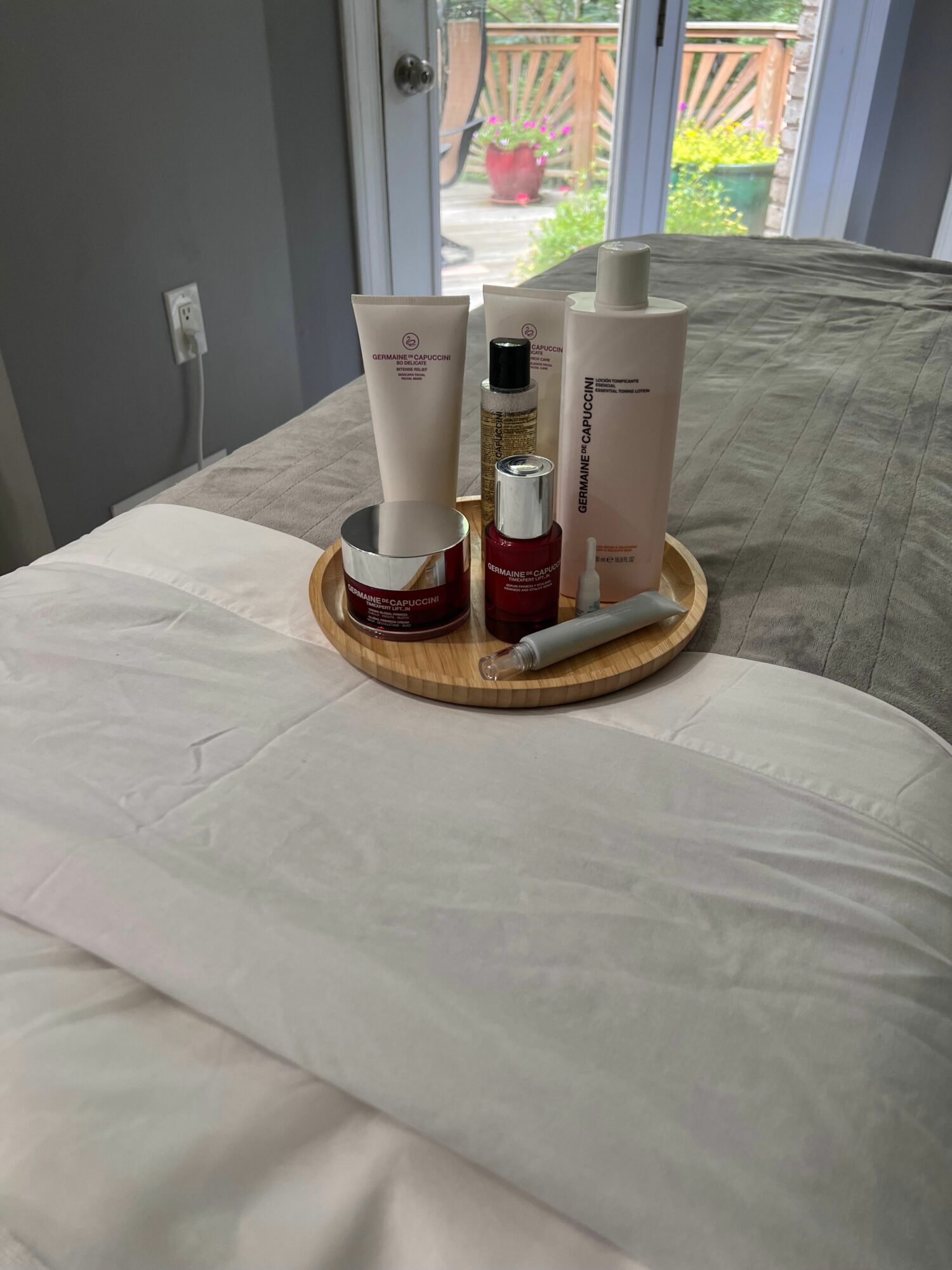Three bottles of skincare products on a round wooden tray on a white bedspread, with a window and outdoor view in the background.