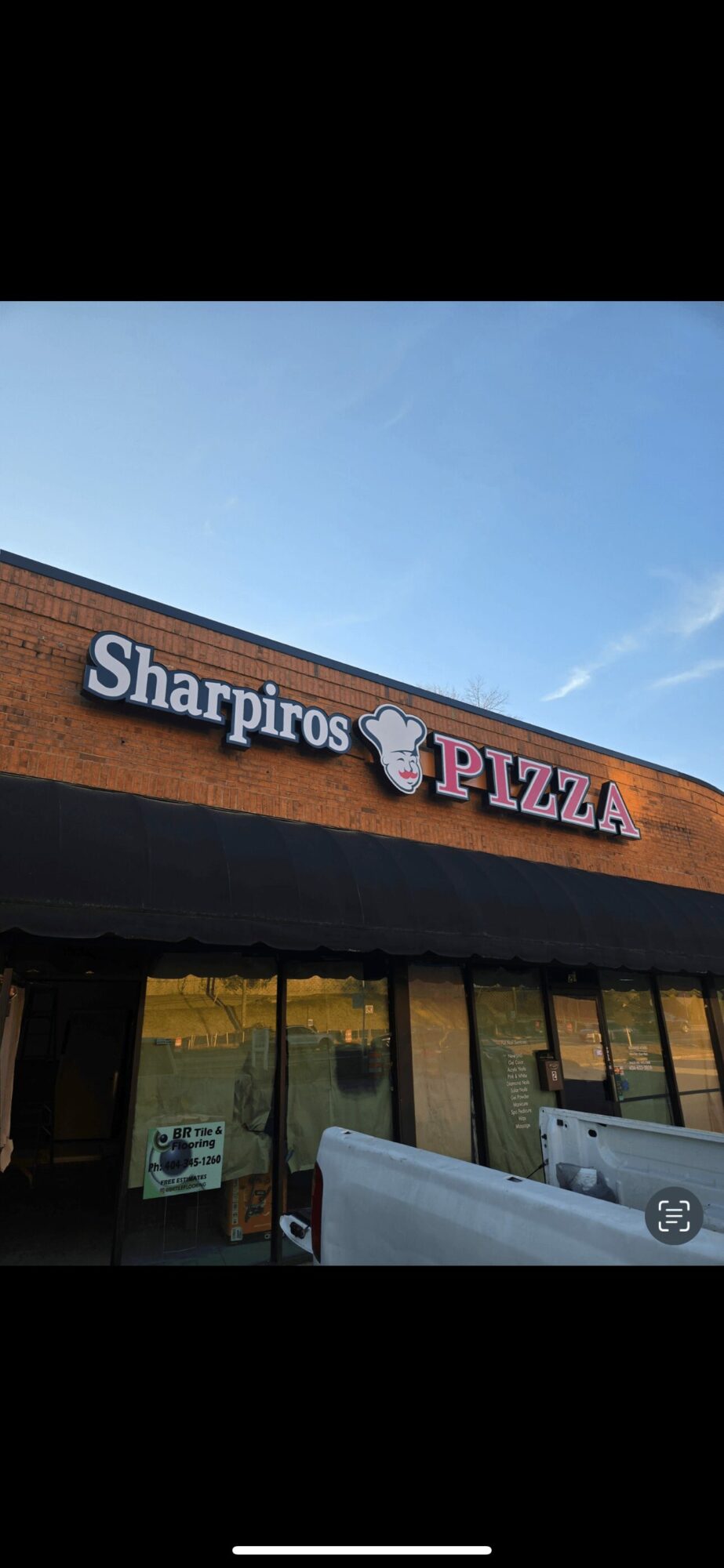 Storefront with sign reading 'Sharpiro PIZZA' and a pig mascot, under a black awning, against a blue sky.