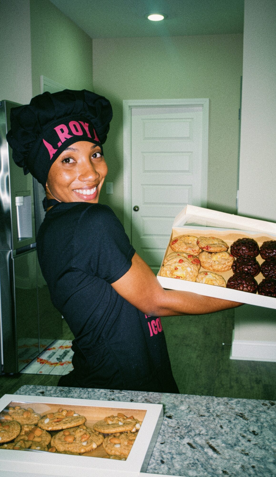 Woman smiling holding a box of cookies in a kitchen, wearing a black headwrap and shirt, with a door in the background.