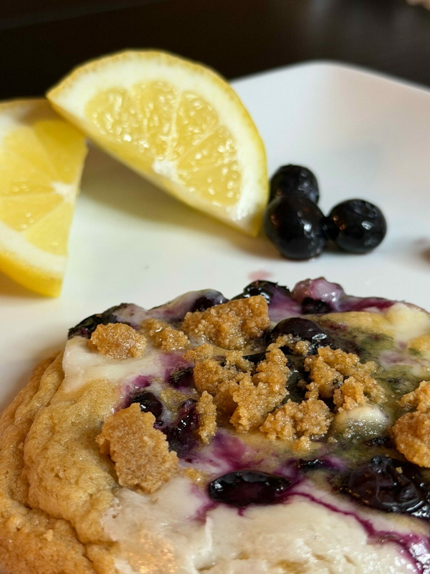 Lemon wedge, blueberries, and a dessert with blueberries, crumble topping, and cream on a white plate.
