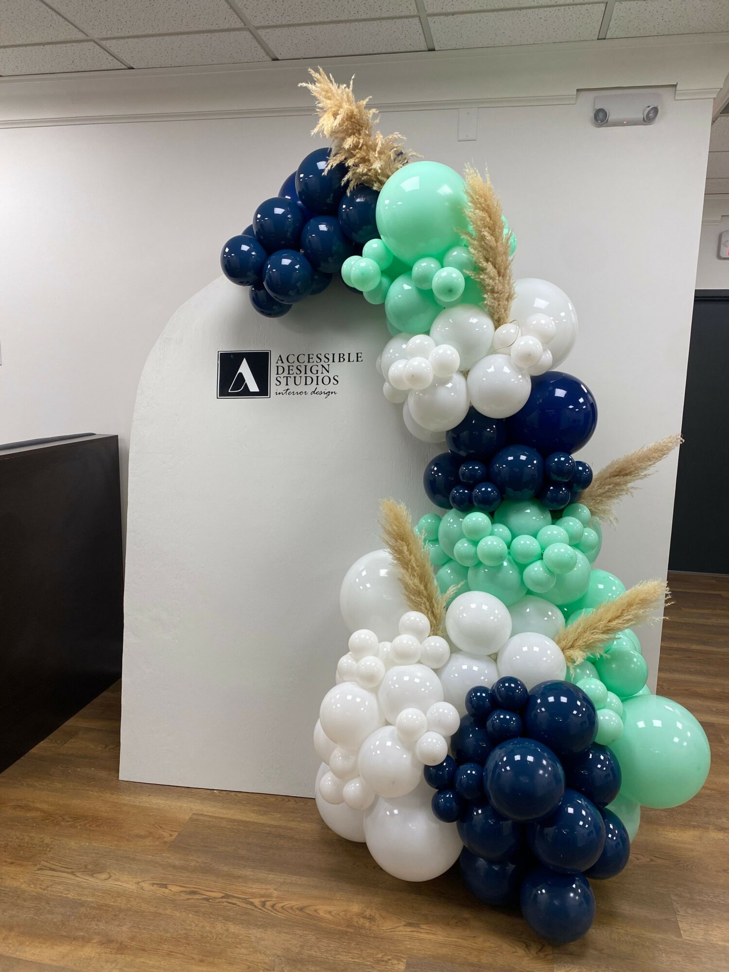 Balloon arrangement with white, navy blue, mint green, and light blue balloons, decorated with wheat stalks, on a wooden floor.