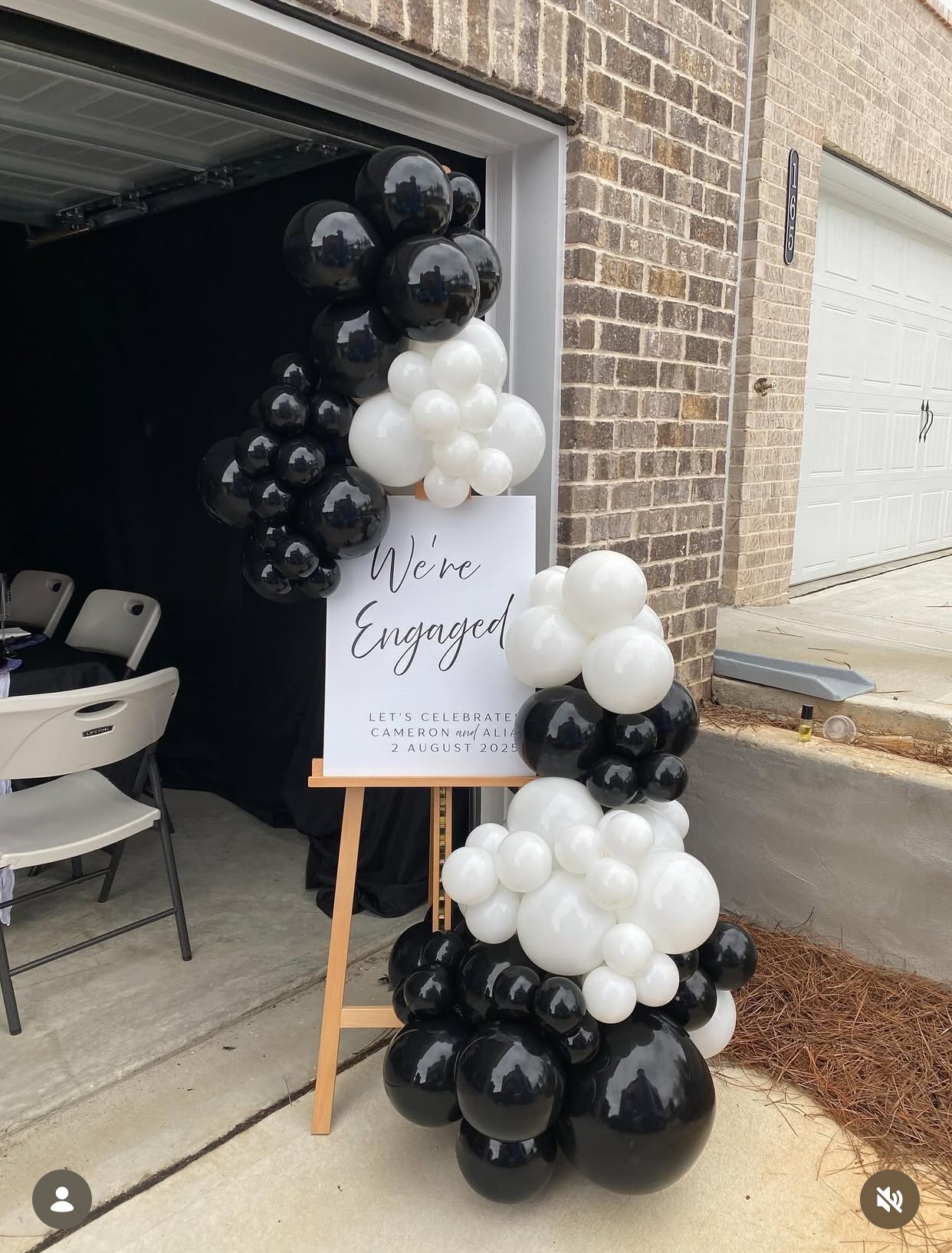 Balloon arrangement with black and white balloons next to a sign that reads 'We're Engaged' outside a garage.