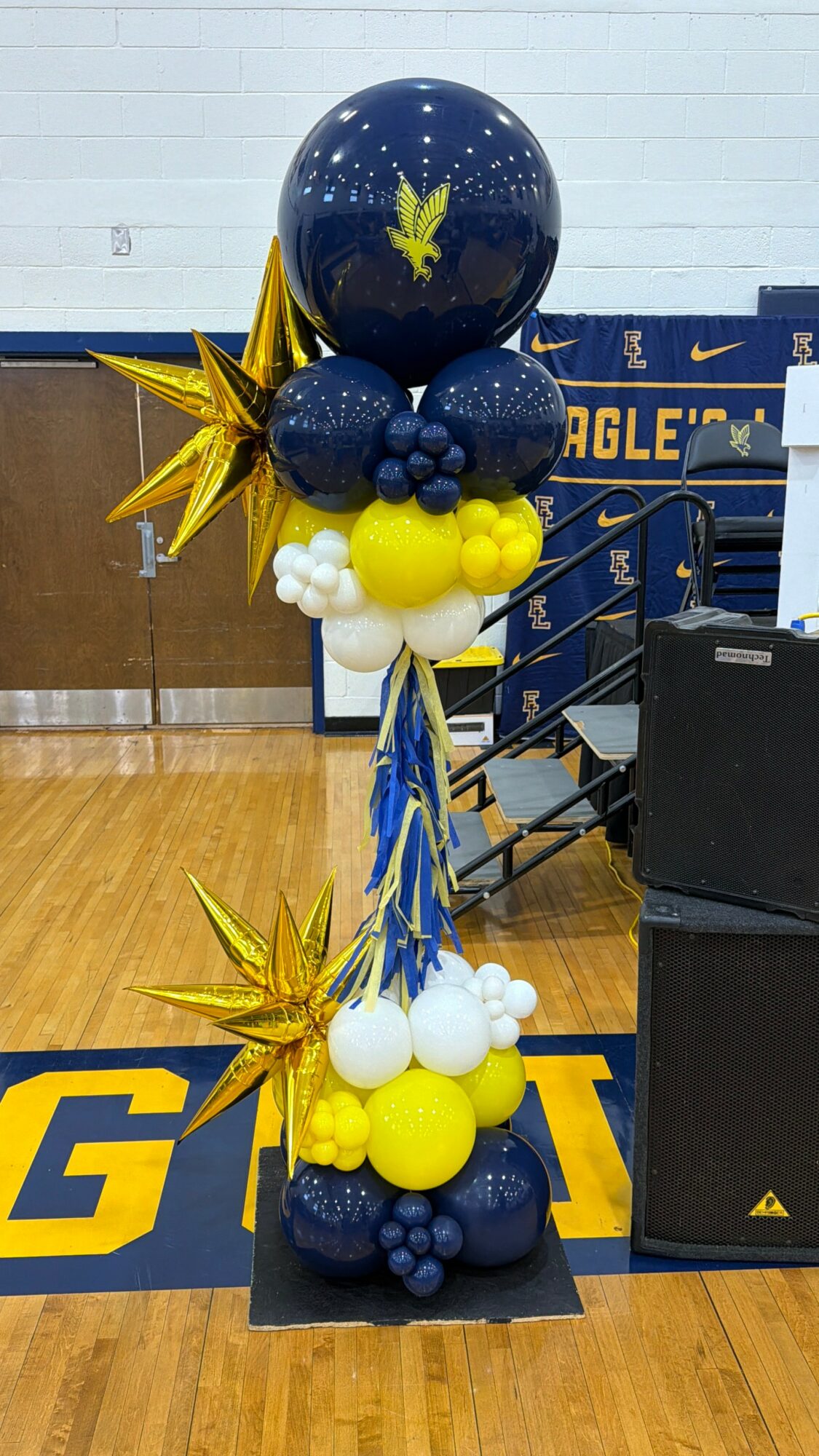 Balloon arrangement with blue, yellow, and white balloons, gold star-shaped balloons, and a large blue balloon with a yellow cannabis leaf, in a gymnasium.