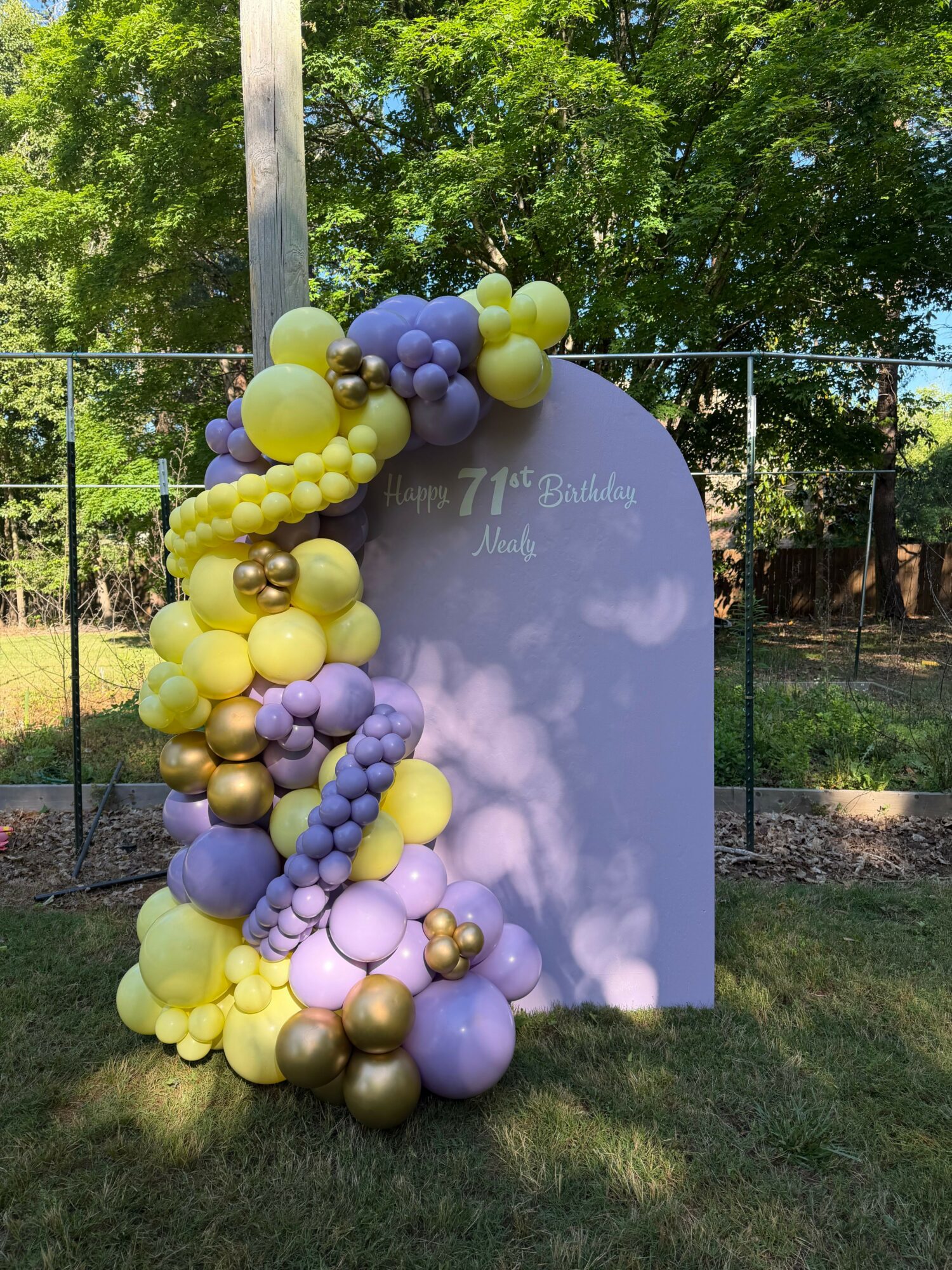 Balloon arrangement with purple, yellow, and gold balloons around a vertical sign on grass, trees in background.