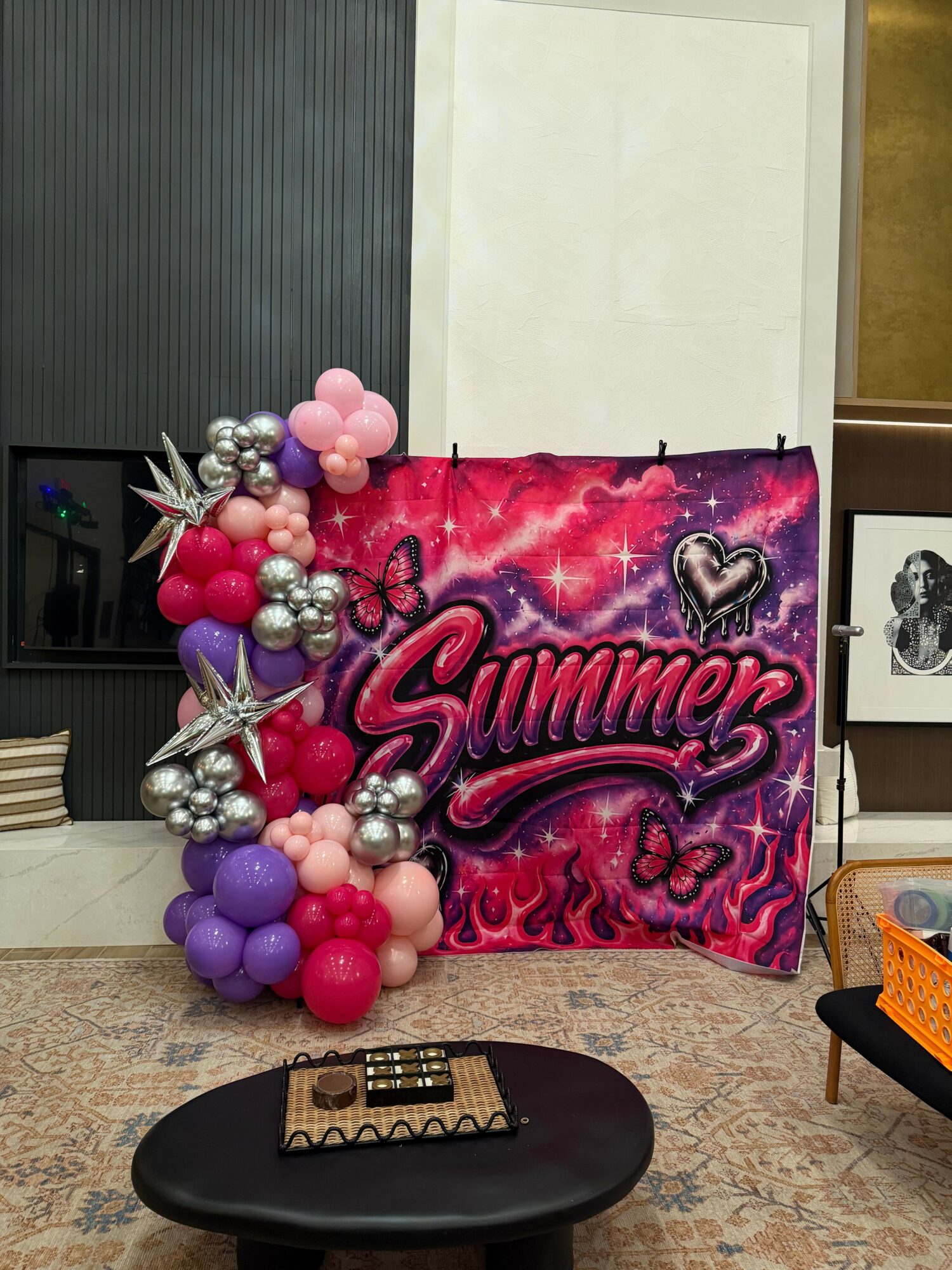 Decorative backdrop with pink, purple, and silver balloons, and a red fabric with pink and black text, in a room.