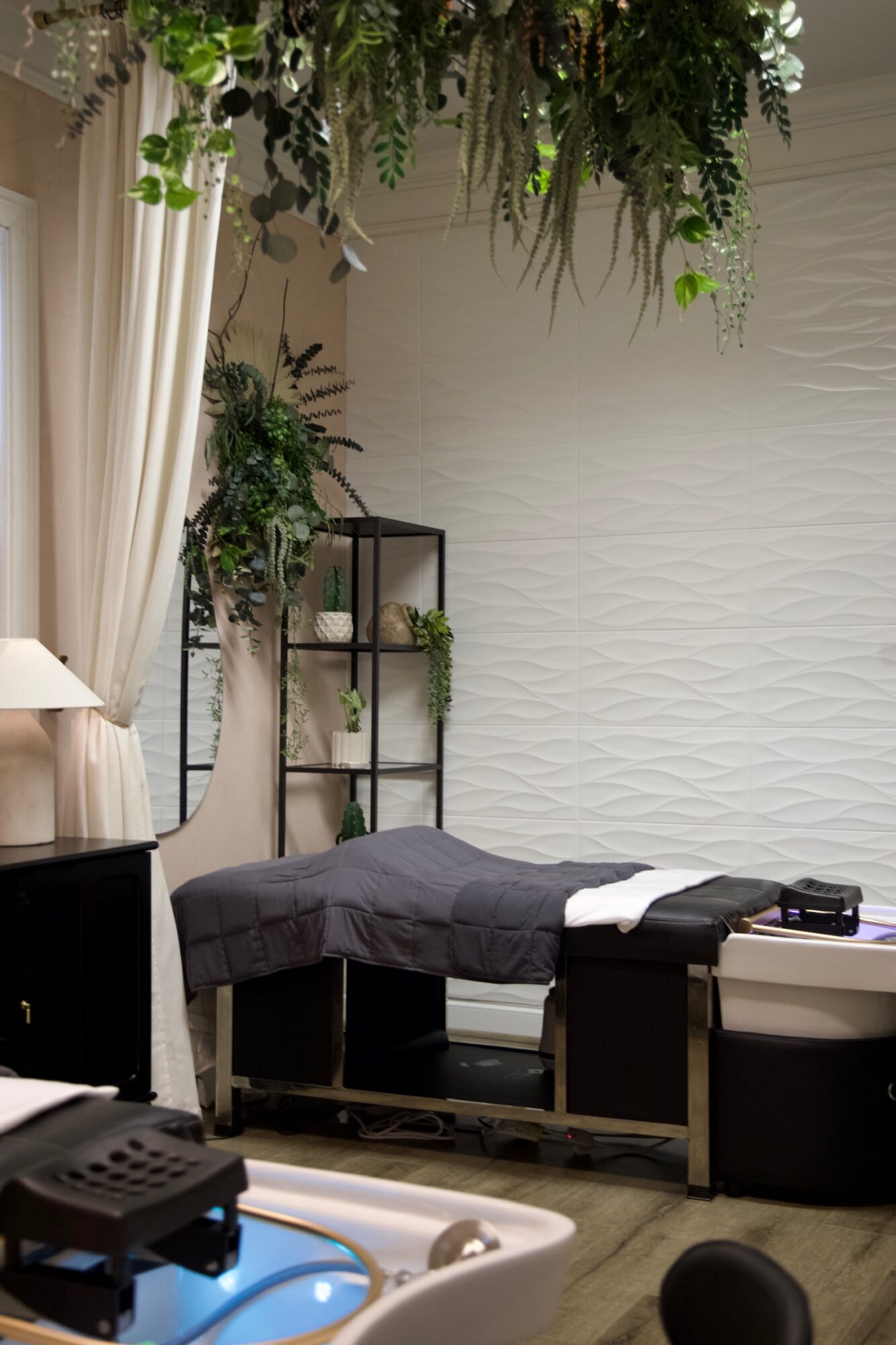 Massage table with dark cover in a room decorated with plants and white textured wall, with a shelf and curtains nearby.