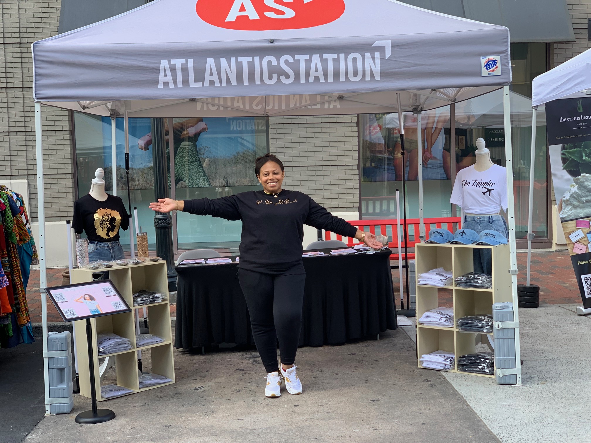 Person standing under a tent with arms outstretched, displaying merchandise and promotional materials, outdoors on a city street.