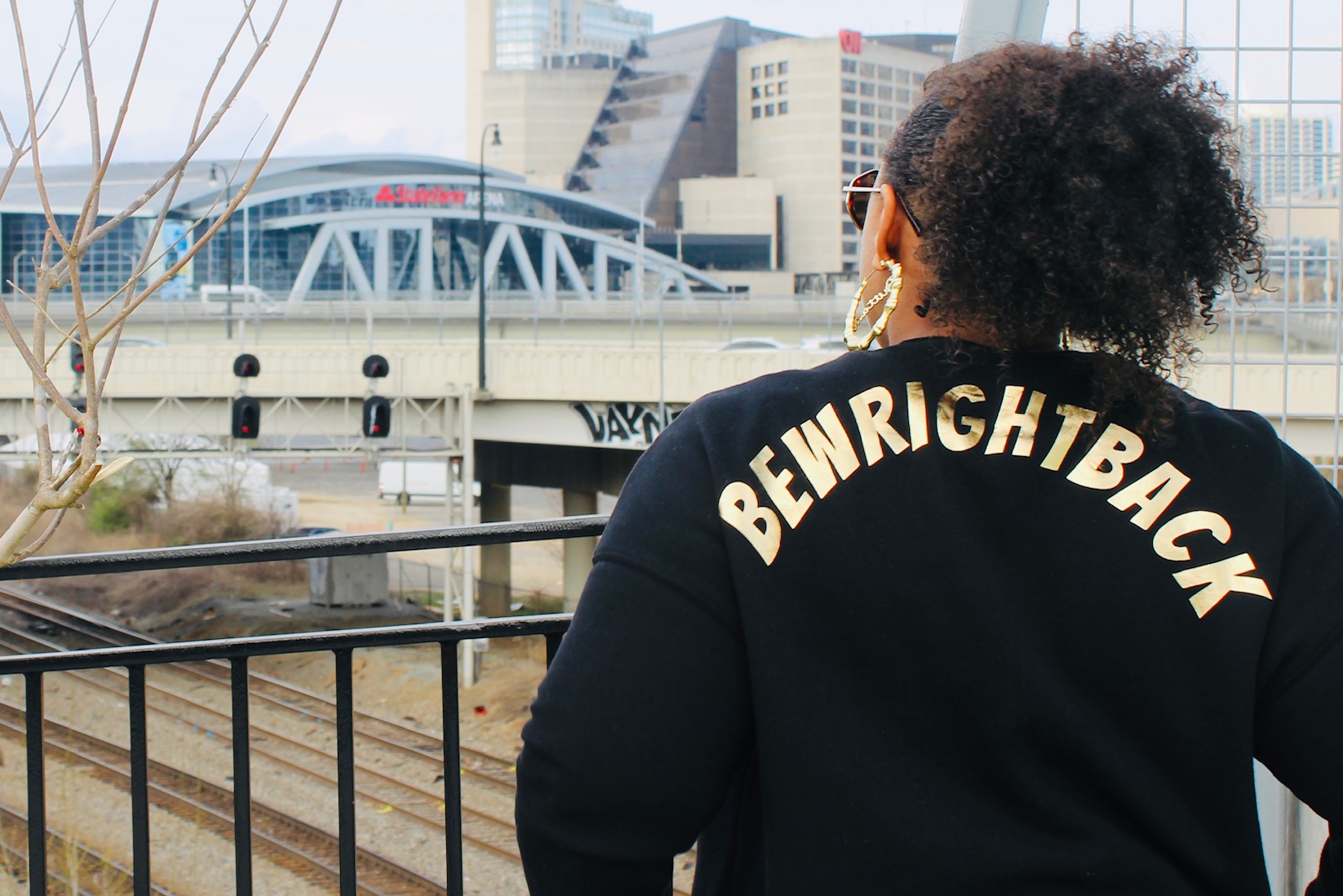 Person with curly hair wearing a black jacket with white text on the back, urban cityscape background.
