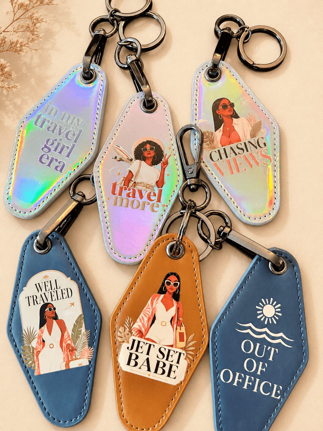 Six colorful keychains with travel and work themes, featuring illustrations of women and text, arranged on a beige surface.