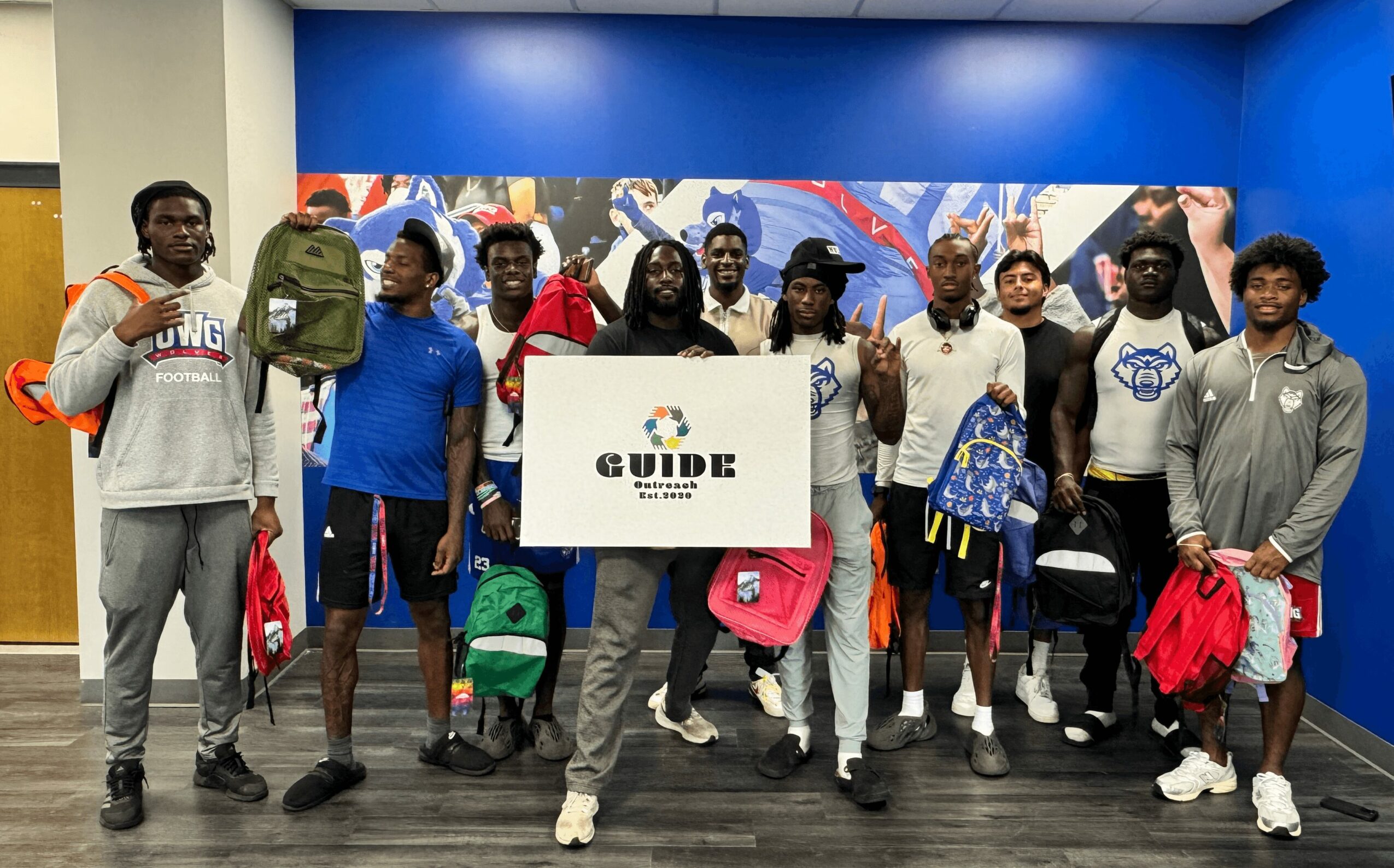 Group of young people standing indoors, holding backpacks and a guide sign, in front of a colorful mural background.