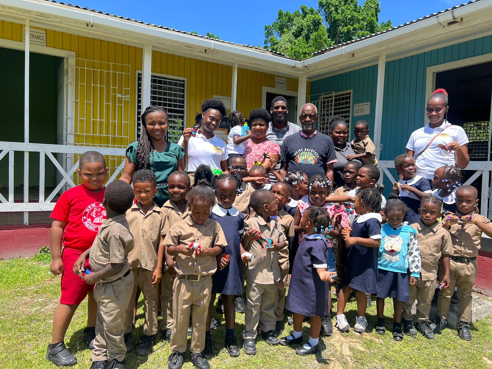 Group of children and adults standing outside colorful buildings, smiling, on a sunny day.